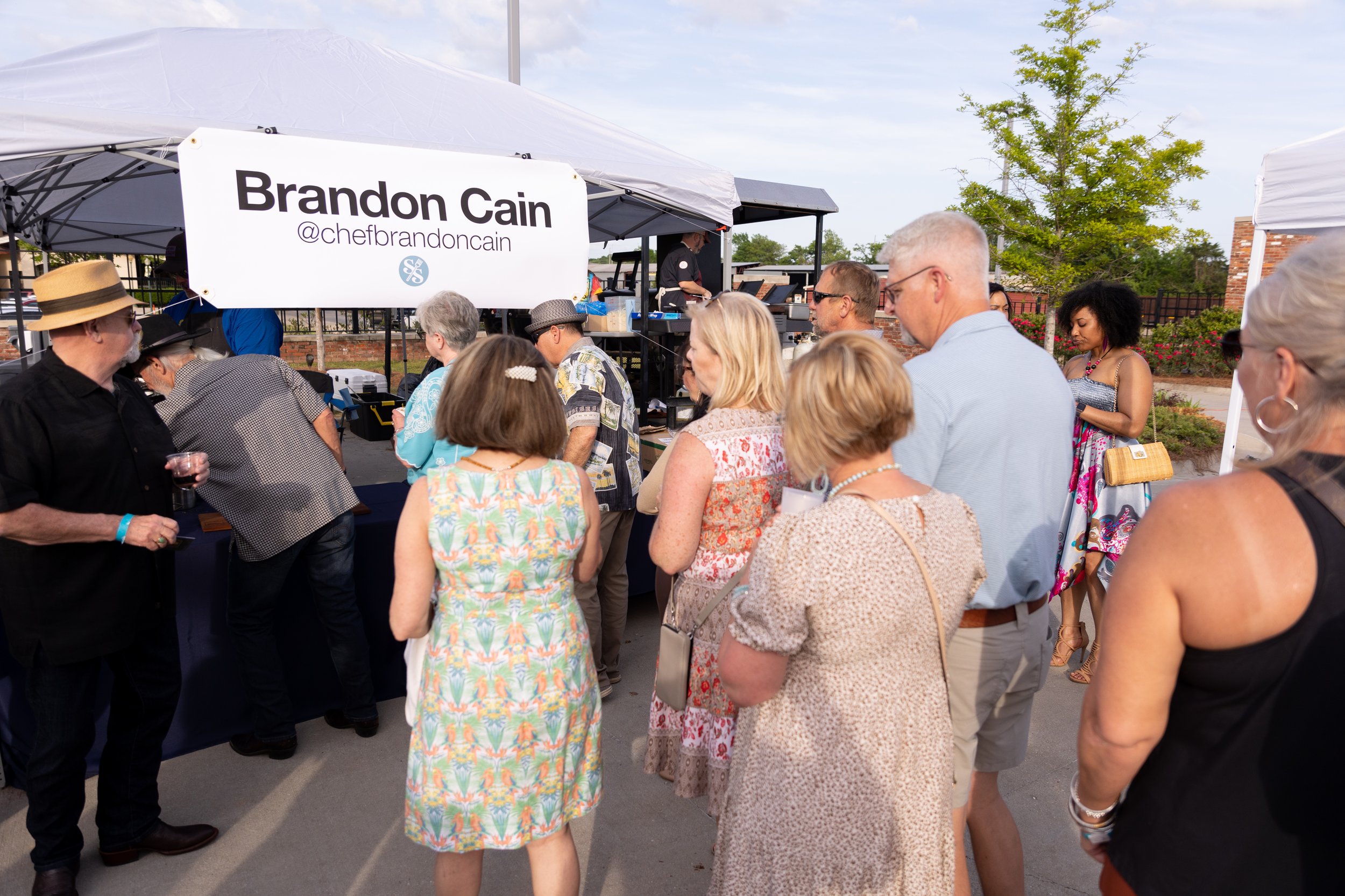 Group of people at an outdoor event in front of a booth with a sign reading 'Brandon Cain @chefbrandoncain' under a tent.