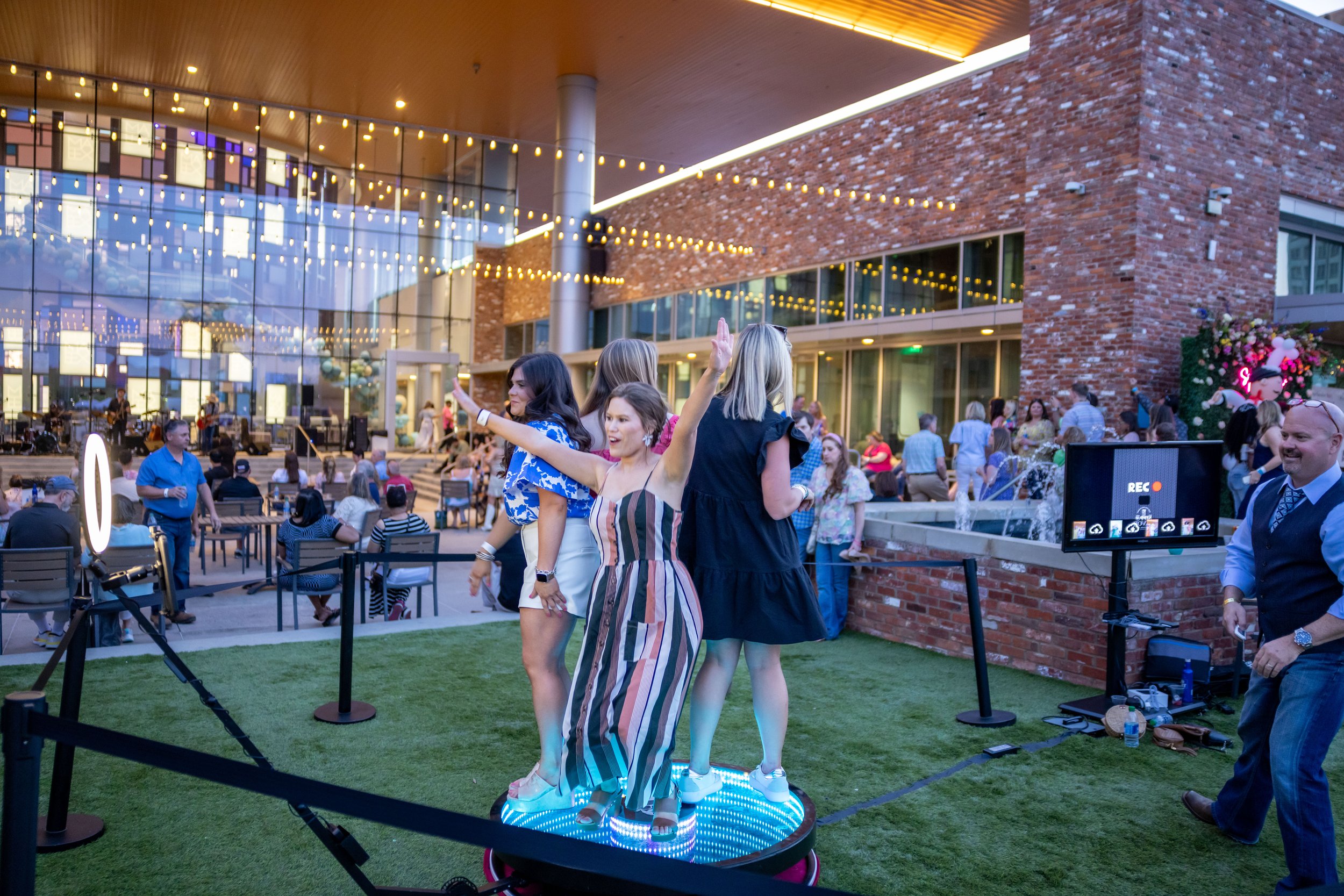 Group of women posing on a 360-degree photo booth platform outdoors, with people sitting in a patio area and string lights above. Brick building in the background.