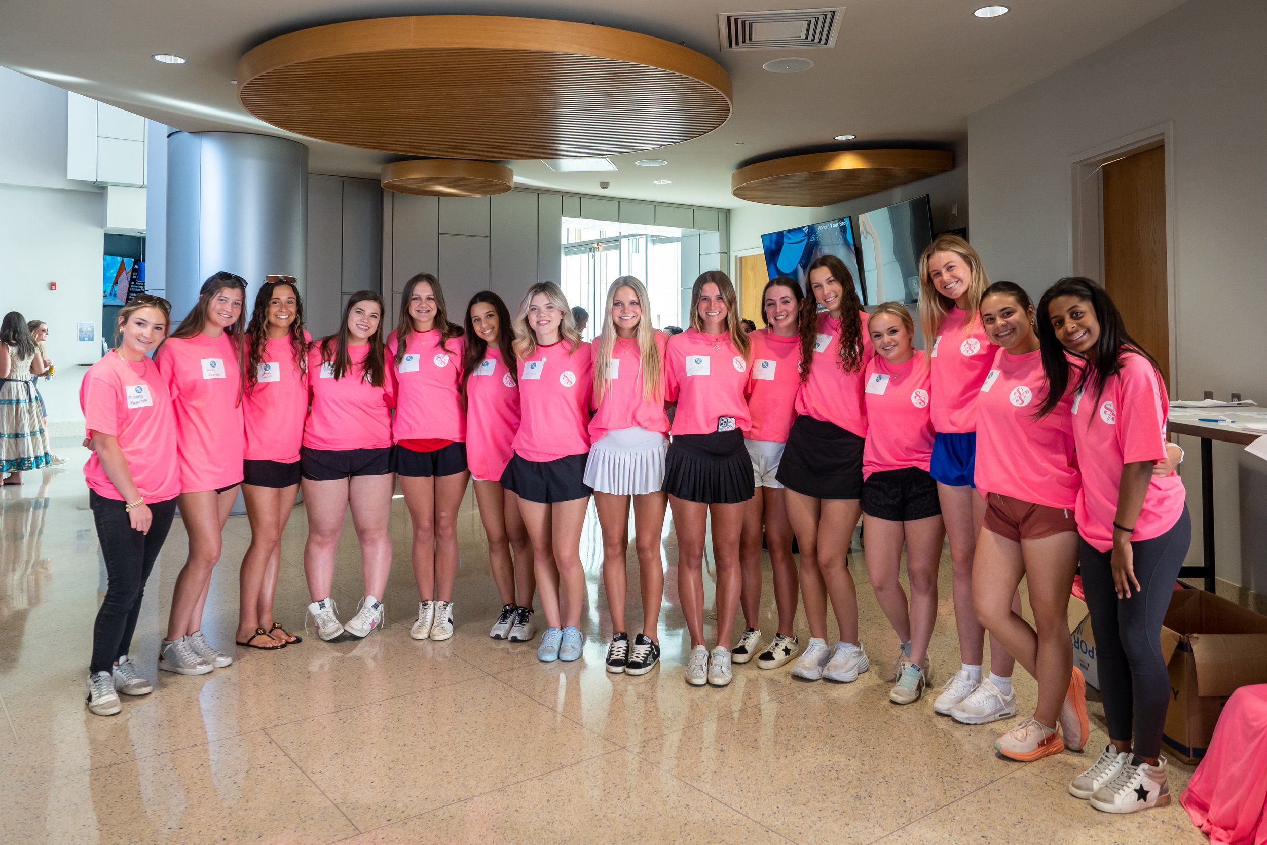 Group of young women wearing matching pink shirts and black or colorful shorts standing together indoors, smiling for the camera.