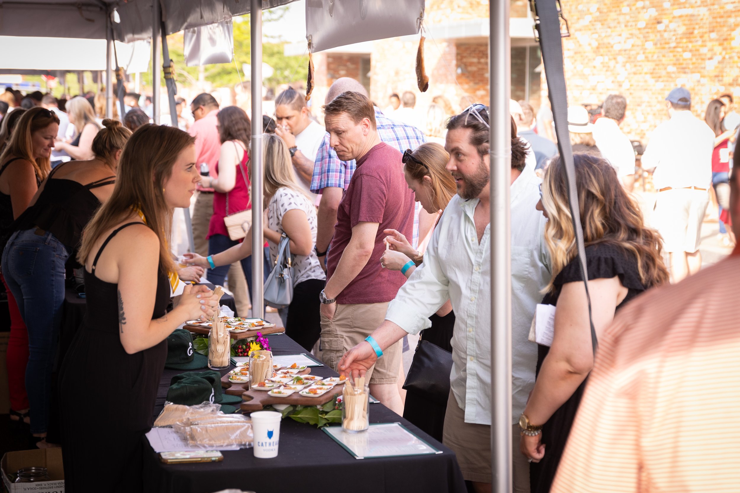 People at an outdoor food festival, sampling dishes from vendor tables under tents.