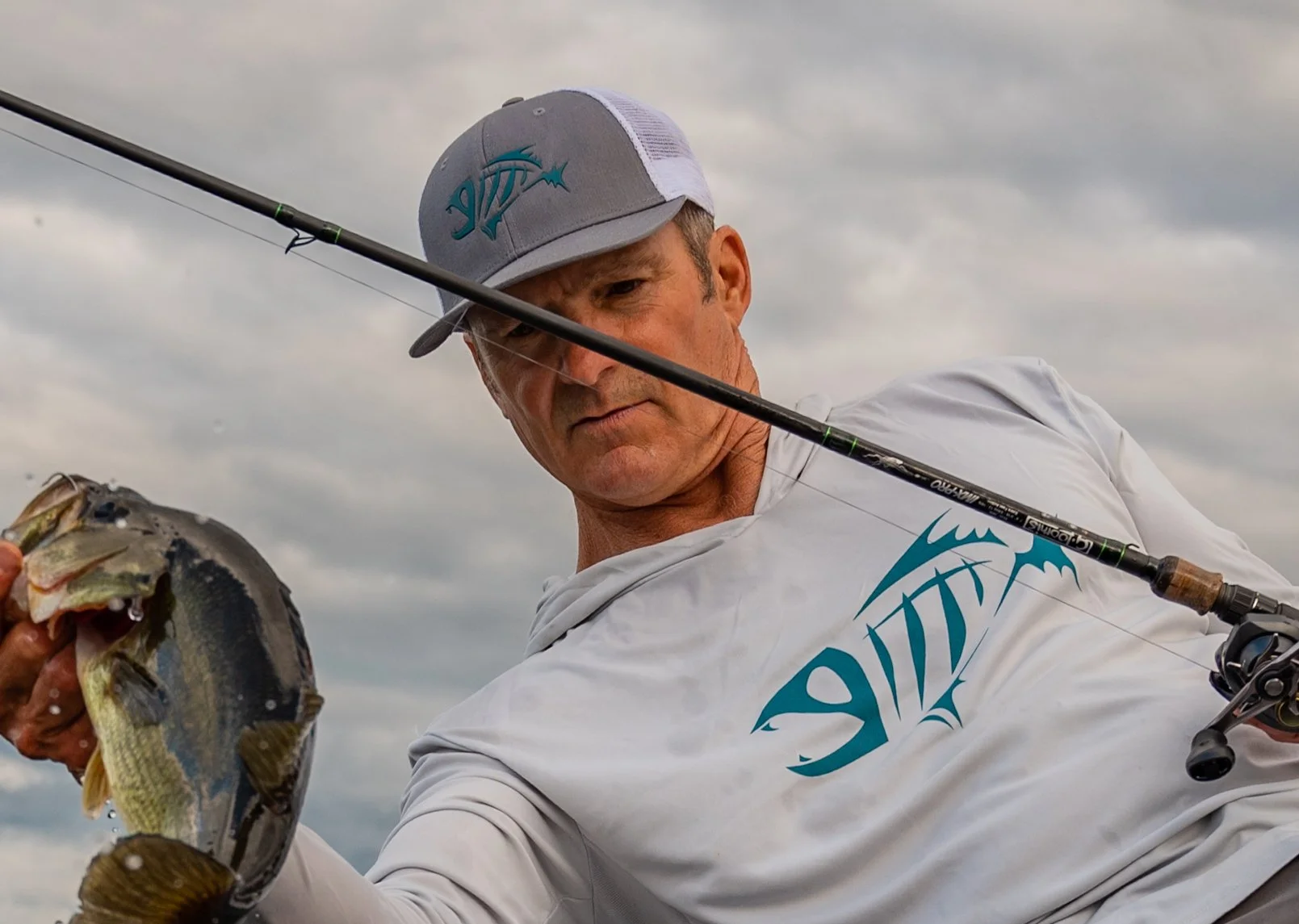 Man holding a fish and fishing rod, wearing a hat and shirt with a fish logo.
