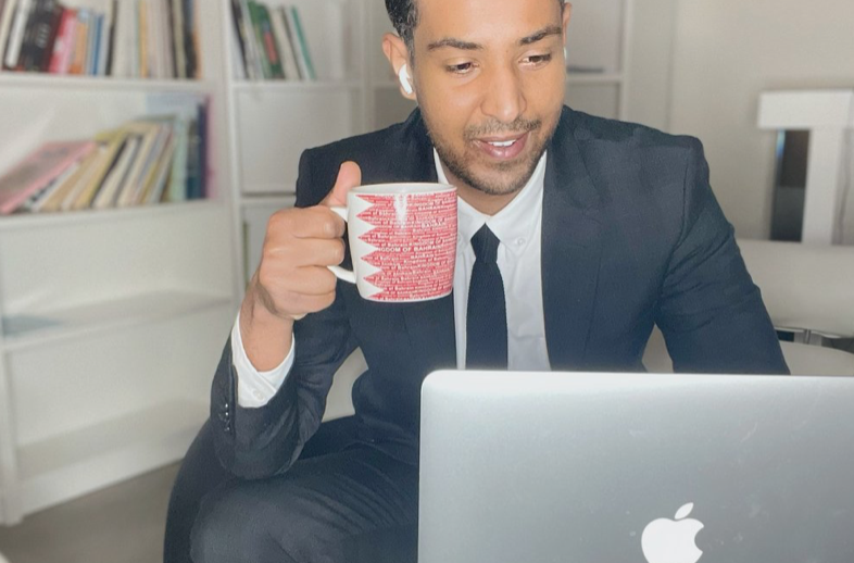 A man in a suit holding a red and white mug, sitting at a desk and looking at a silver MacBook laptop in a modern office or home setting.