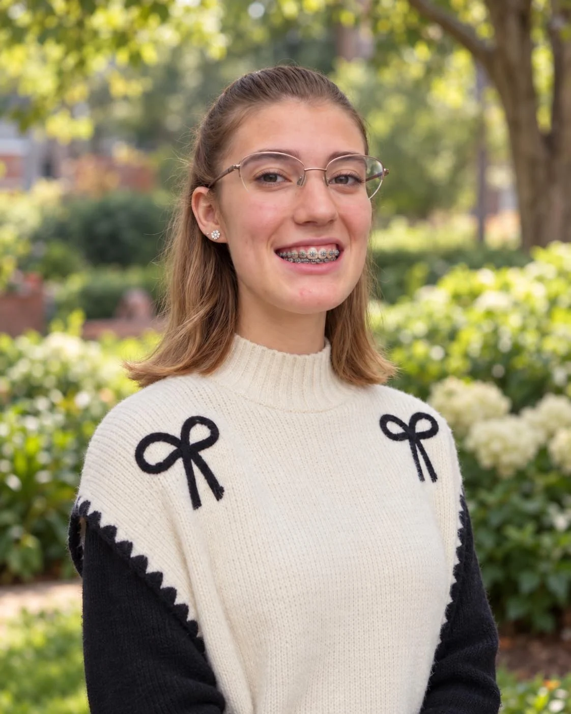 Young woman with glasses and a ponytail, smiling outdoors in front of green foliage.