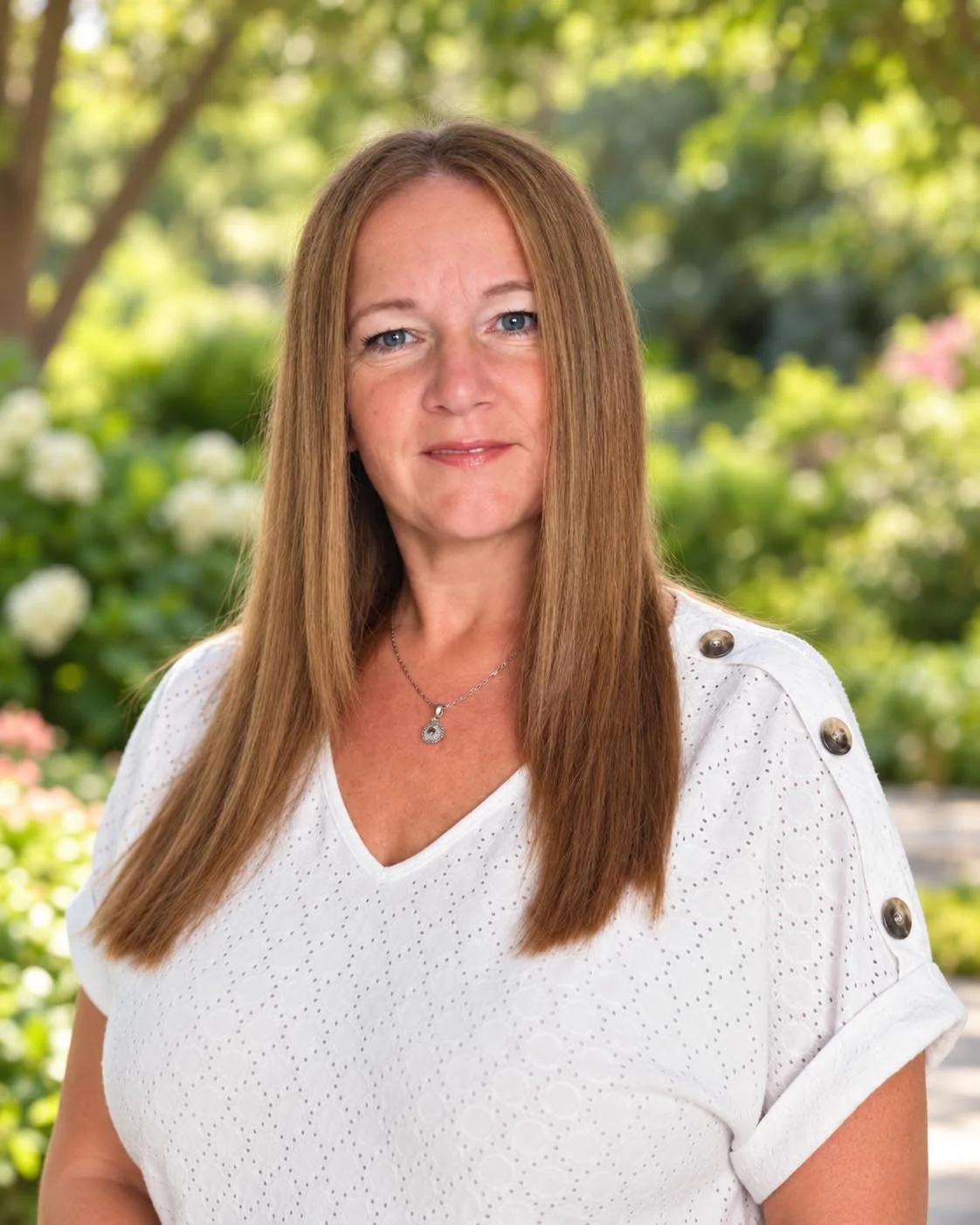 A woman with straight, shoulder-length red hair, wearing a white blazer over a black top, standing outdoors with green foliage in the background.