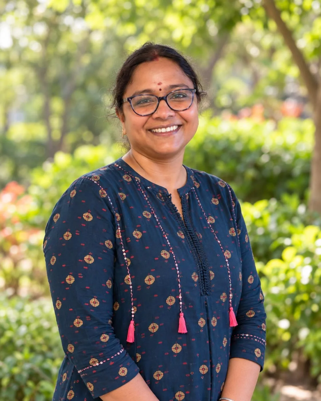 A woman smiling outdoors with green foliage in the background.