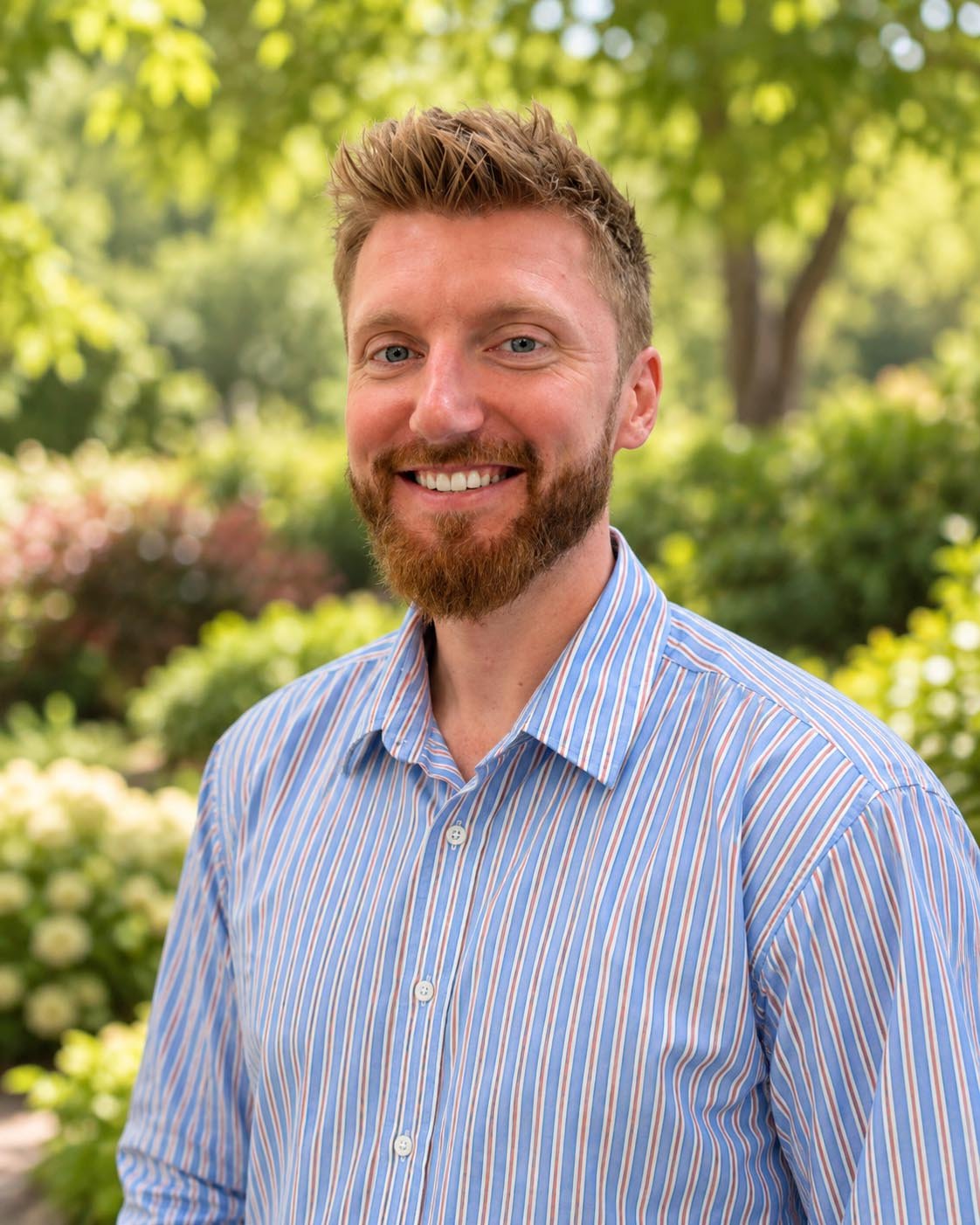 Portrait of a smiling man with a beard and short hair, wearing a plaid shirt, standing outdoors in front of green foliage.