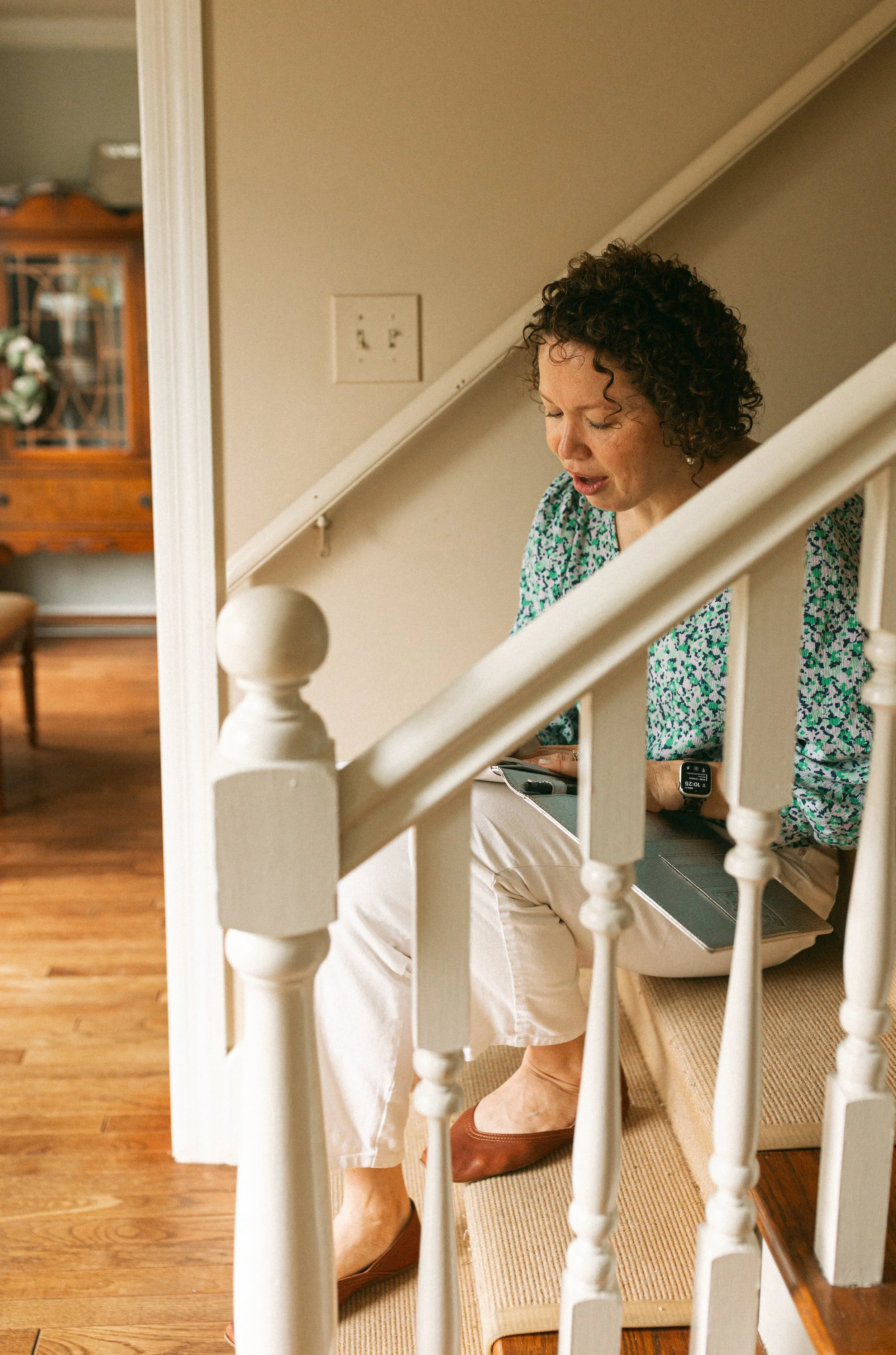 Woman with curly hair sitting on a stairway, looking down, wearing a green patterned blouse, beige pants, and brown shoes.