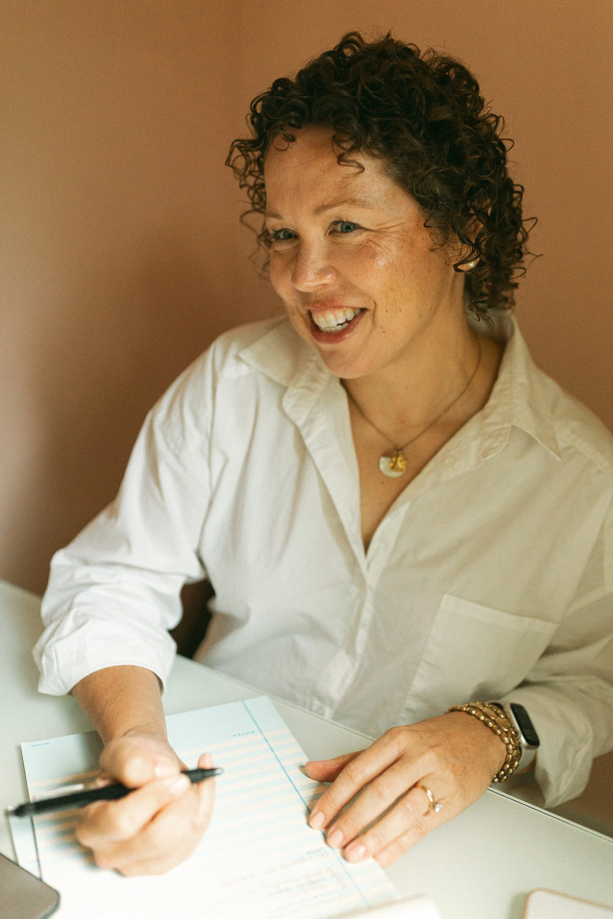A woman with curly hair wearing a white shirt, gold jewelry, and a smartwatch, sitting at a desk, writing in a lined notebook with a black pen.