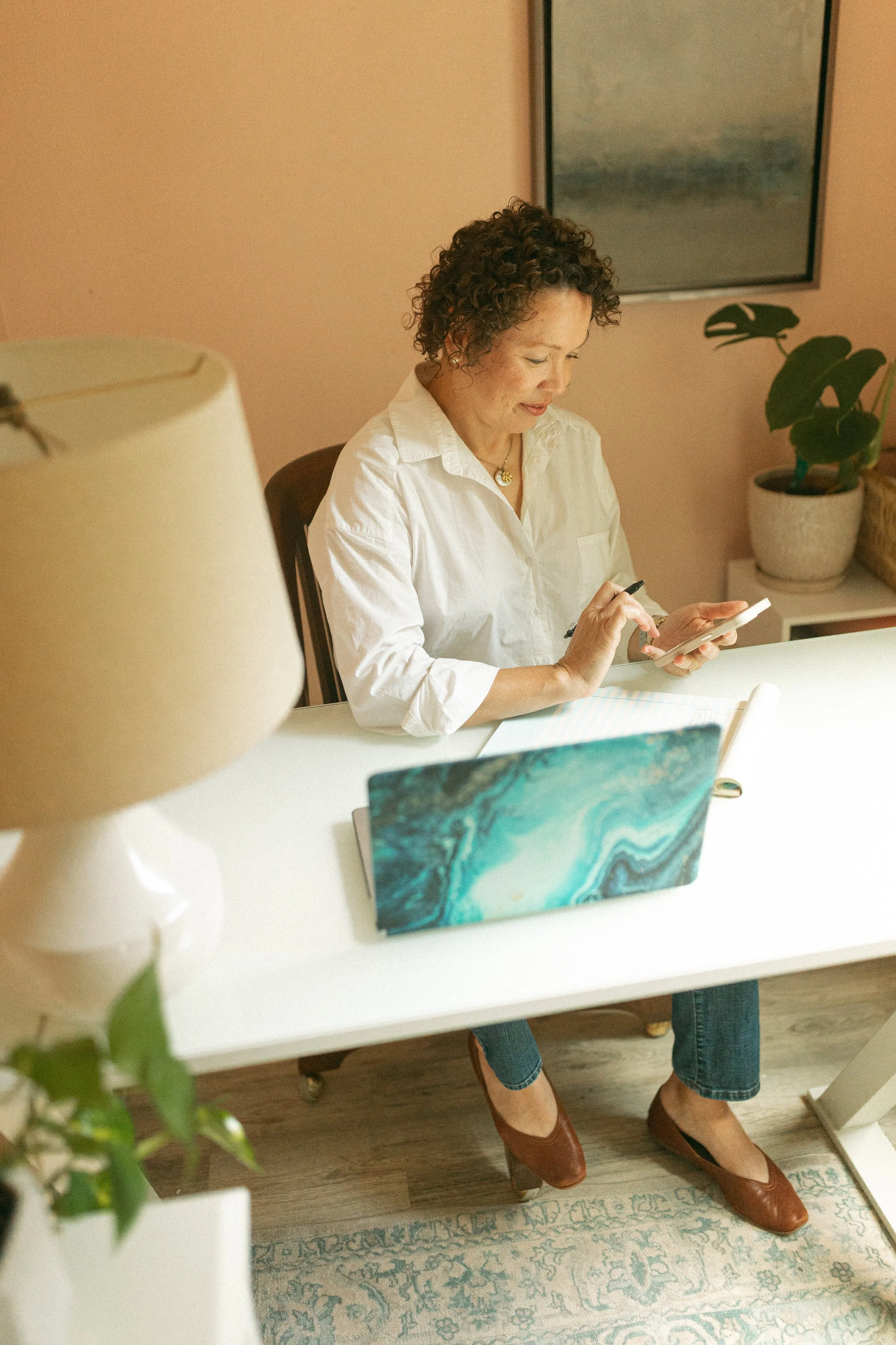 Woman sitting at a desk using a phone while holding a pen in front of a pad of paper and a laptop