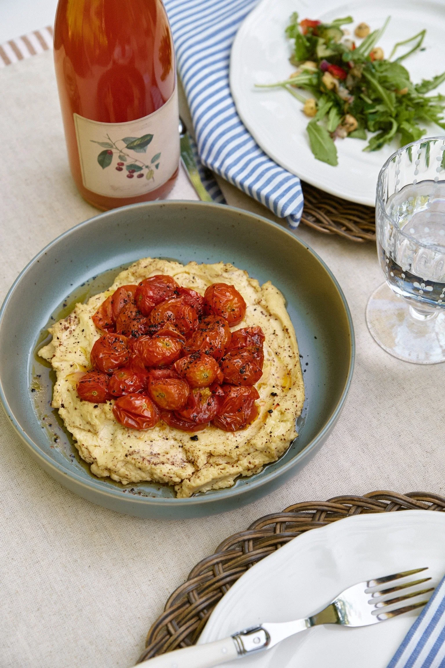 roasted cherry tomatoes over a bowl of hummus, salad on white plate in background, with a blue and white stripe napkins. bottle of cider and a glass of water in background.