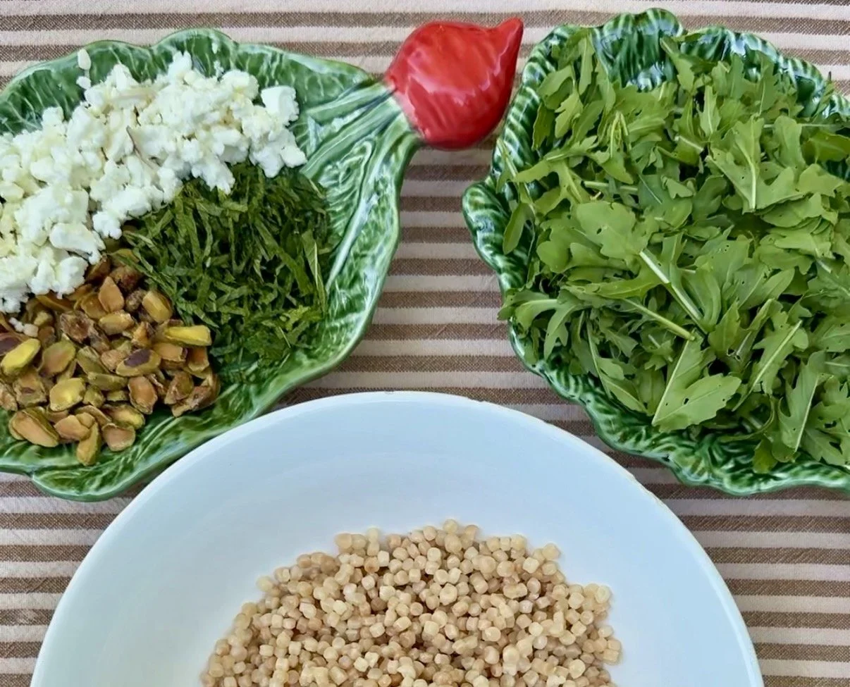 Bowls with pearl couscous, arugula, mint, pistachios, and feta on a striped tablecloth
