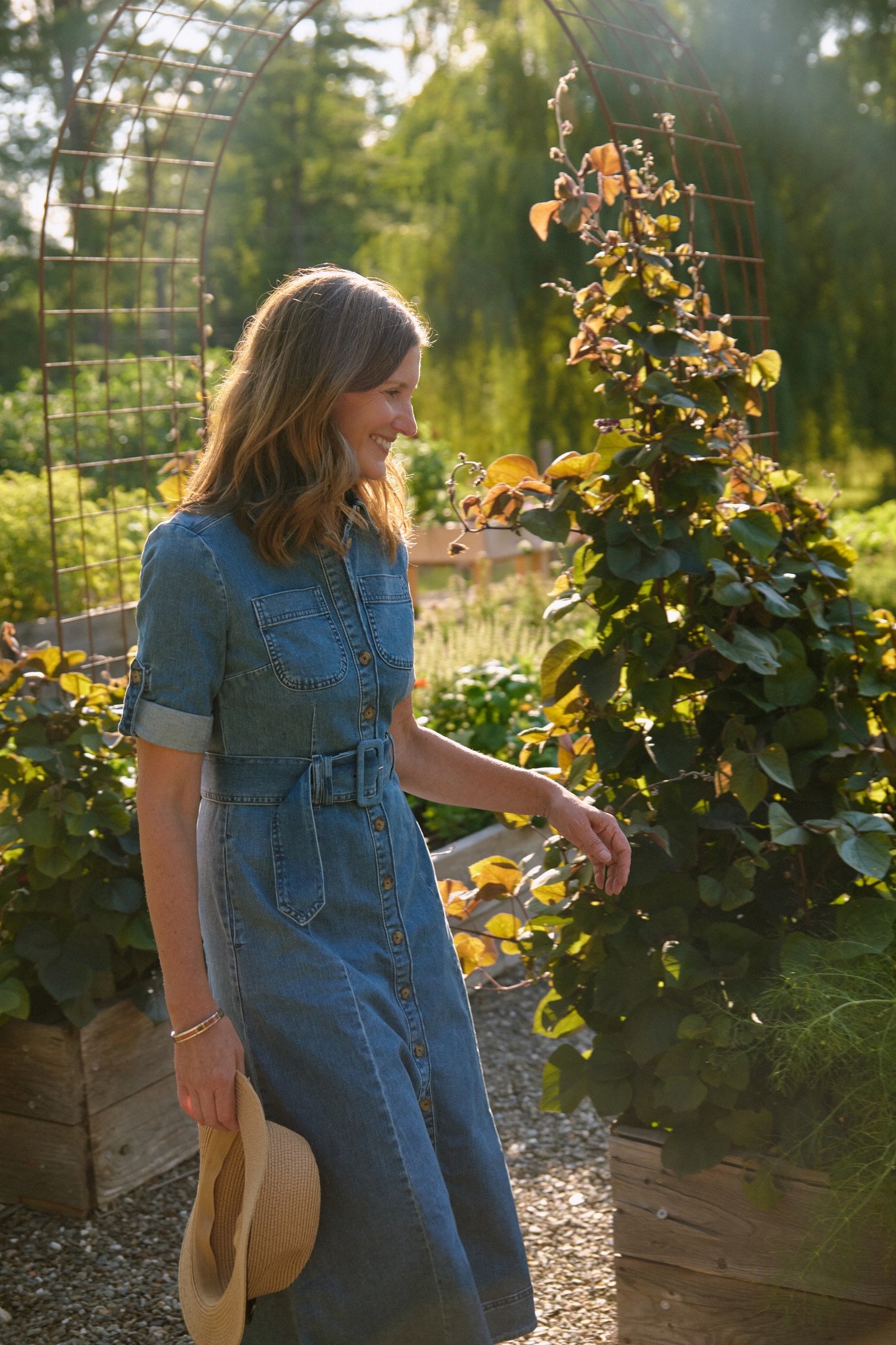 Woman in denim dress walking in a vegetable garden, past an arch trellis planted with vining beans