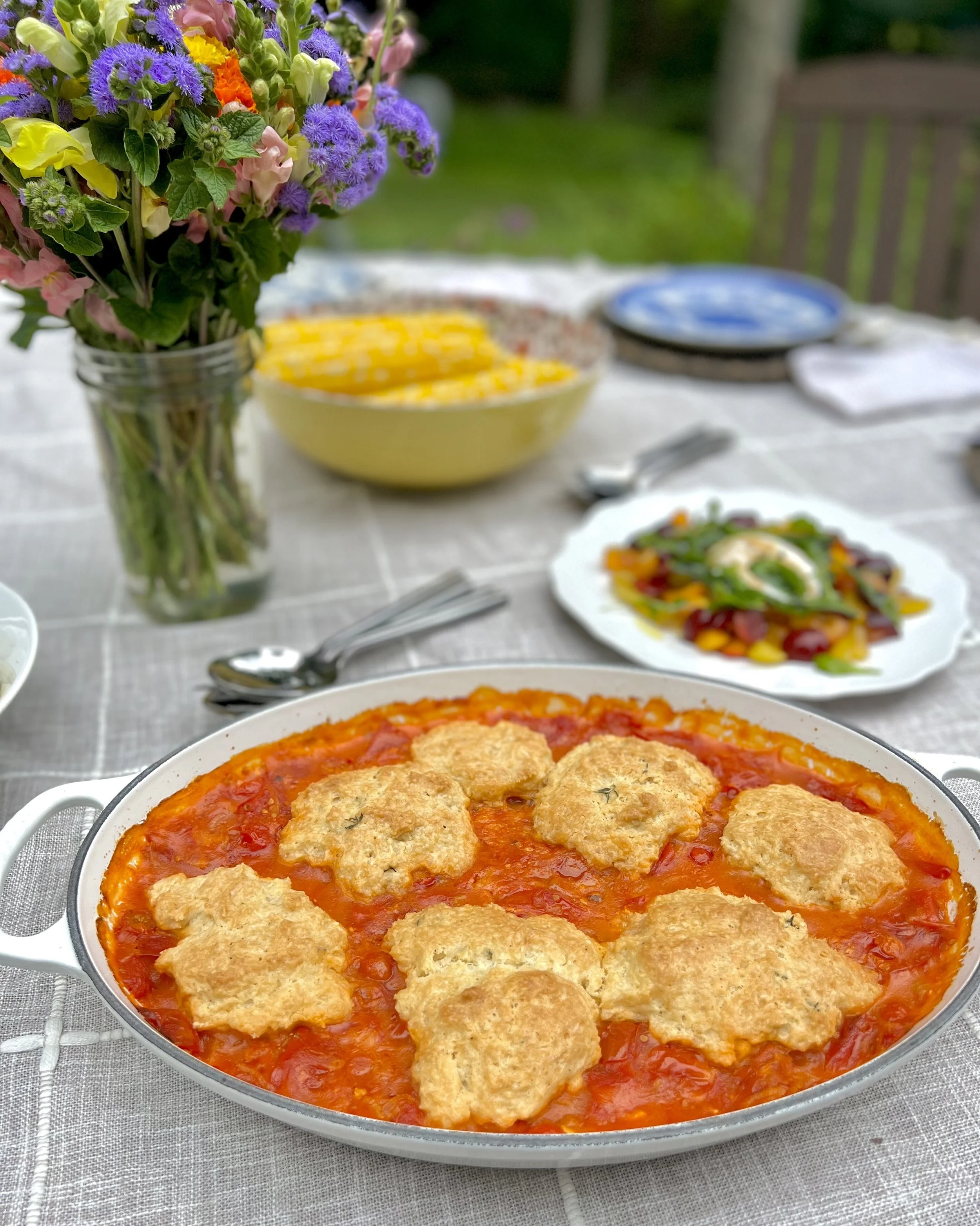 white serving dish with red tomato sauce and biscuit topping set on an outdoor dining table with a grey and white windowpane tablecloth. flowers, salad, corn in the background.