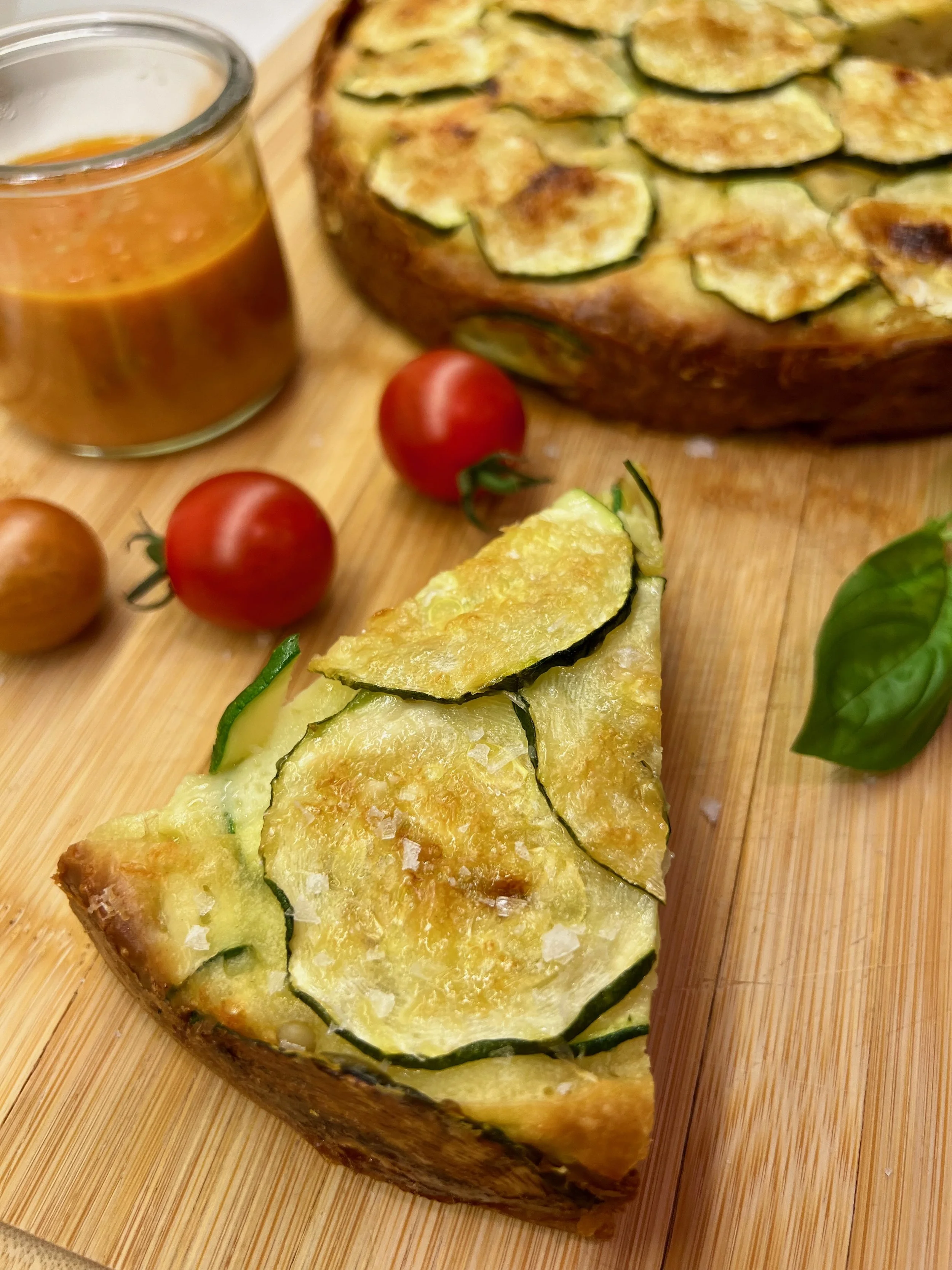 Slice of tart with slices of zucchini in the front, cherry tomatoes and full tart in the background, along with a tomato dipping sauce