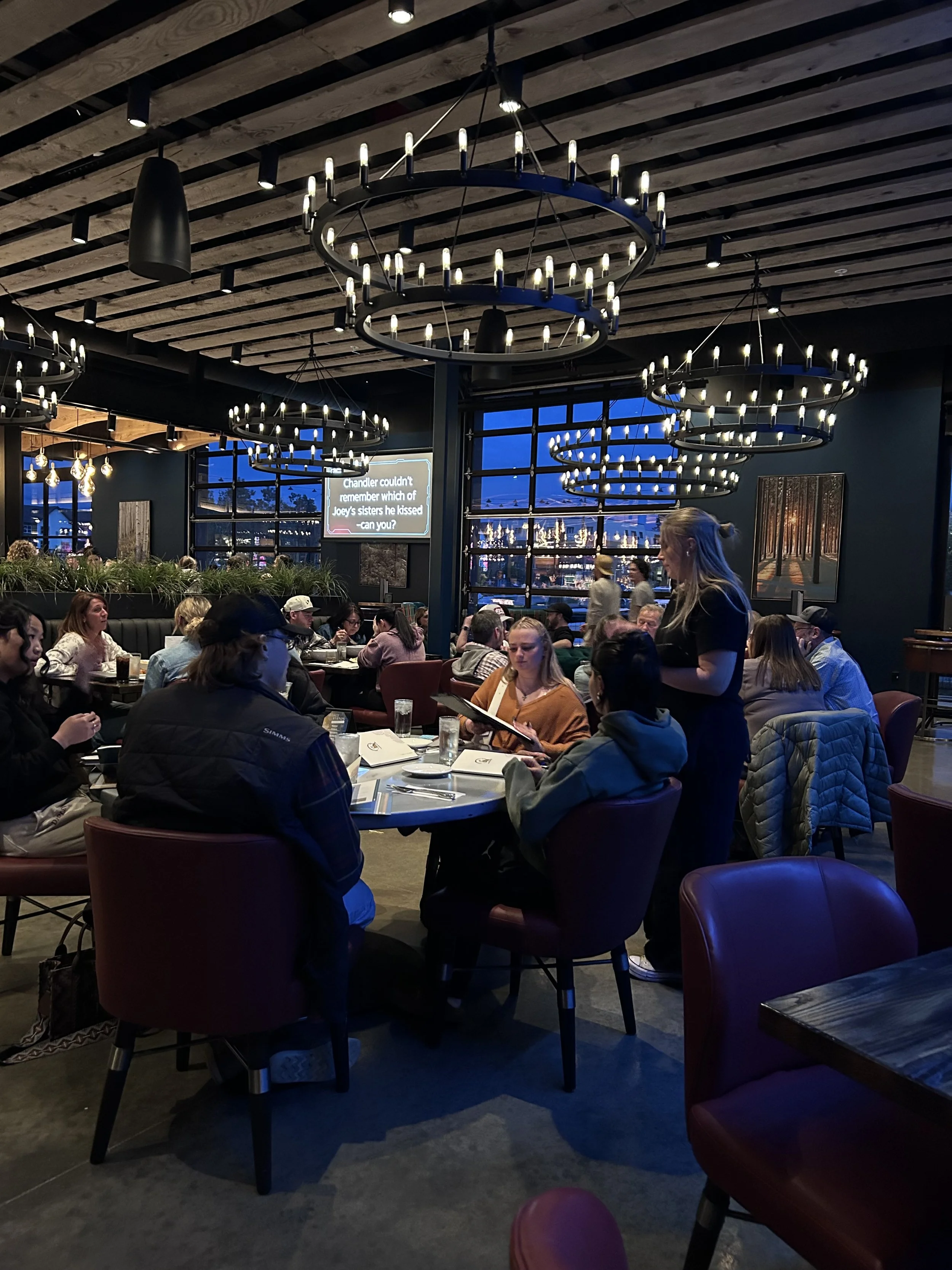 Restaurant filled with people playing trivia in a night time setting with chandelier lighting fixtures overhead.
