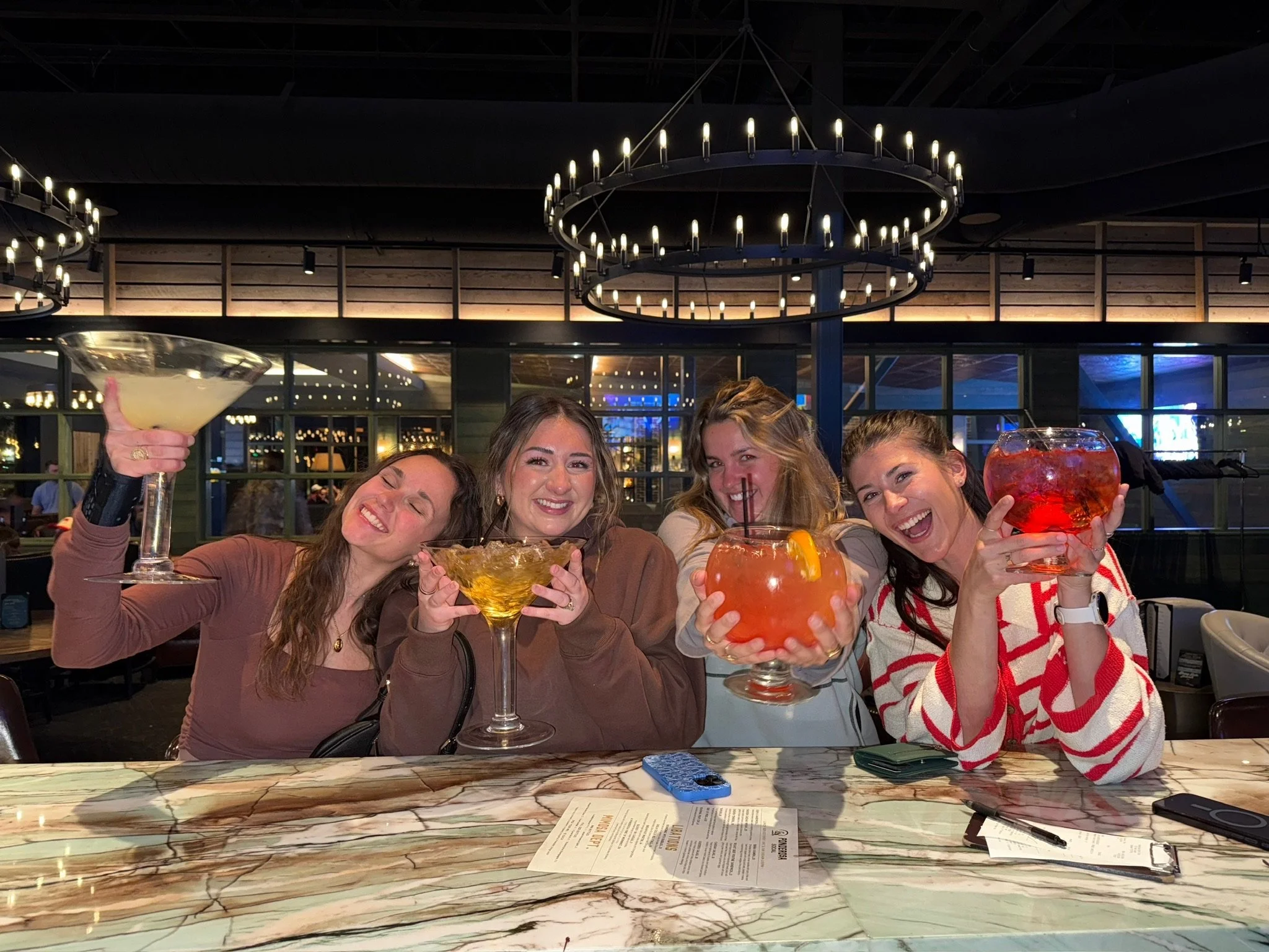 Group of 4 ladies at the bar holding up their cocktail drinks, laughing and smiling.