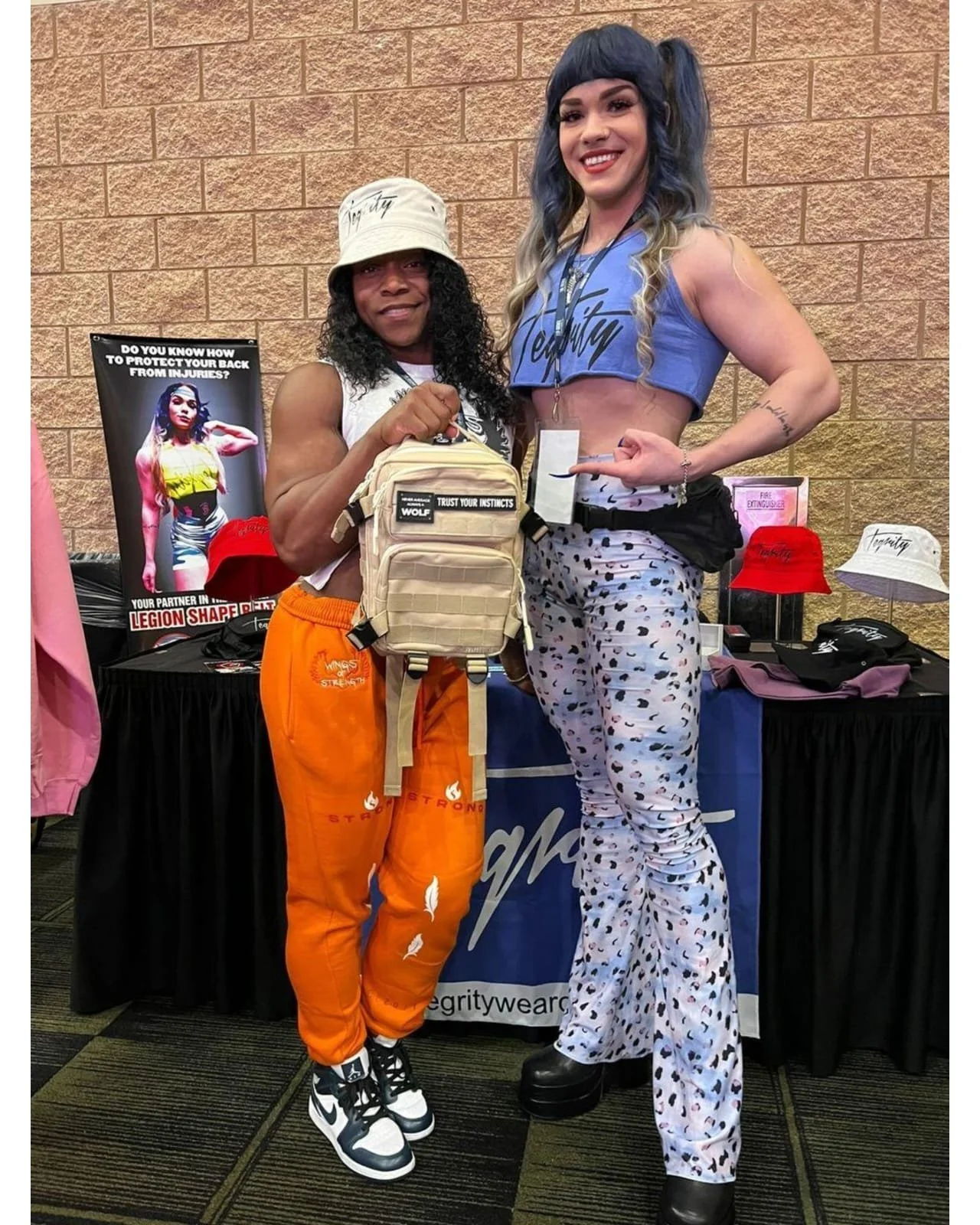 Two women standing together at a booth in a convention or expo. One is wearing orange pants, a beige hat, and holding a tan backpack. The other is dressed in a blue crop top and patterned pants, pointing at her with a smile. Behind them are caps, shi