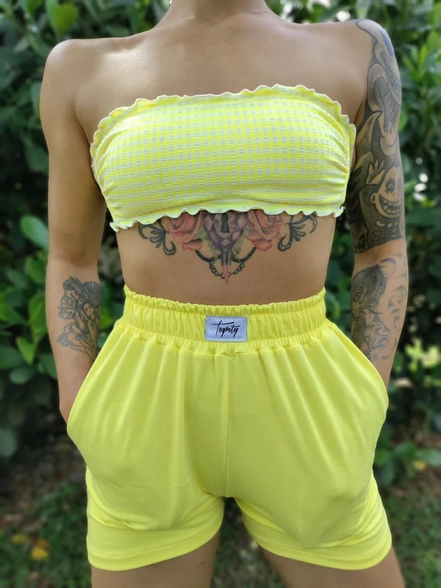 Person wearing a yellow checkered bandeau top and matching yellow shorts with tattoos on arms and a large tattoo on the chest, standing outdoors with green foliage in the background.
