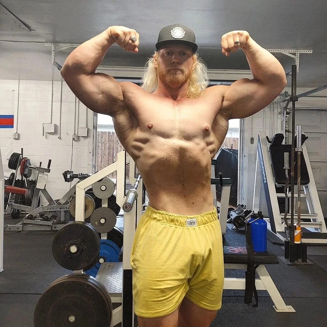A shirtless man with long blond hair, wearing a black cap and yellow shorts, flexing his biceps in a gym.