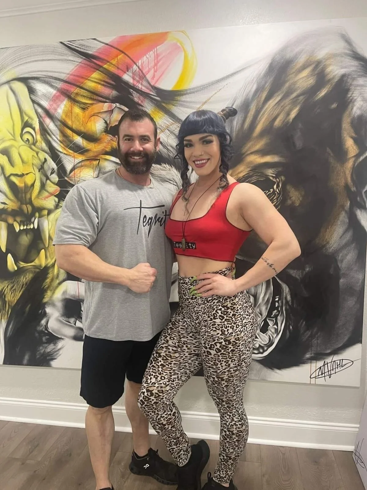 A man with a beard wearing a gray T-shirt and black shorts standing next to a woman with black hair styled in bangs wearing a red crop top and leopard print pants, both smiling and posing in front of a large, vibrant painting of lions.