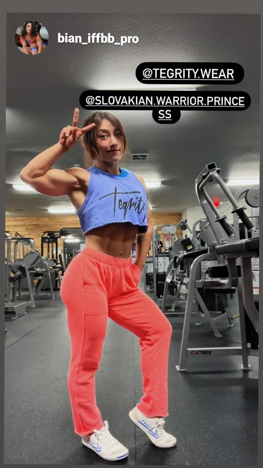 A woman in a gym flexing her arm and smiling at the camera. She is wearing a blue crop top, red sweatpants, and white sneakers. The gym has various exercise machines in the background.