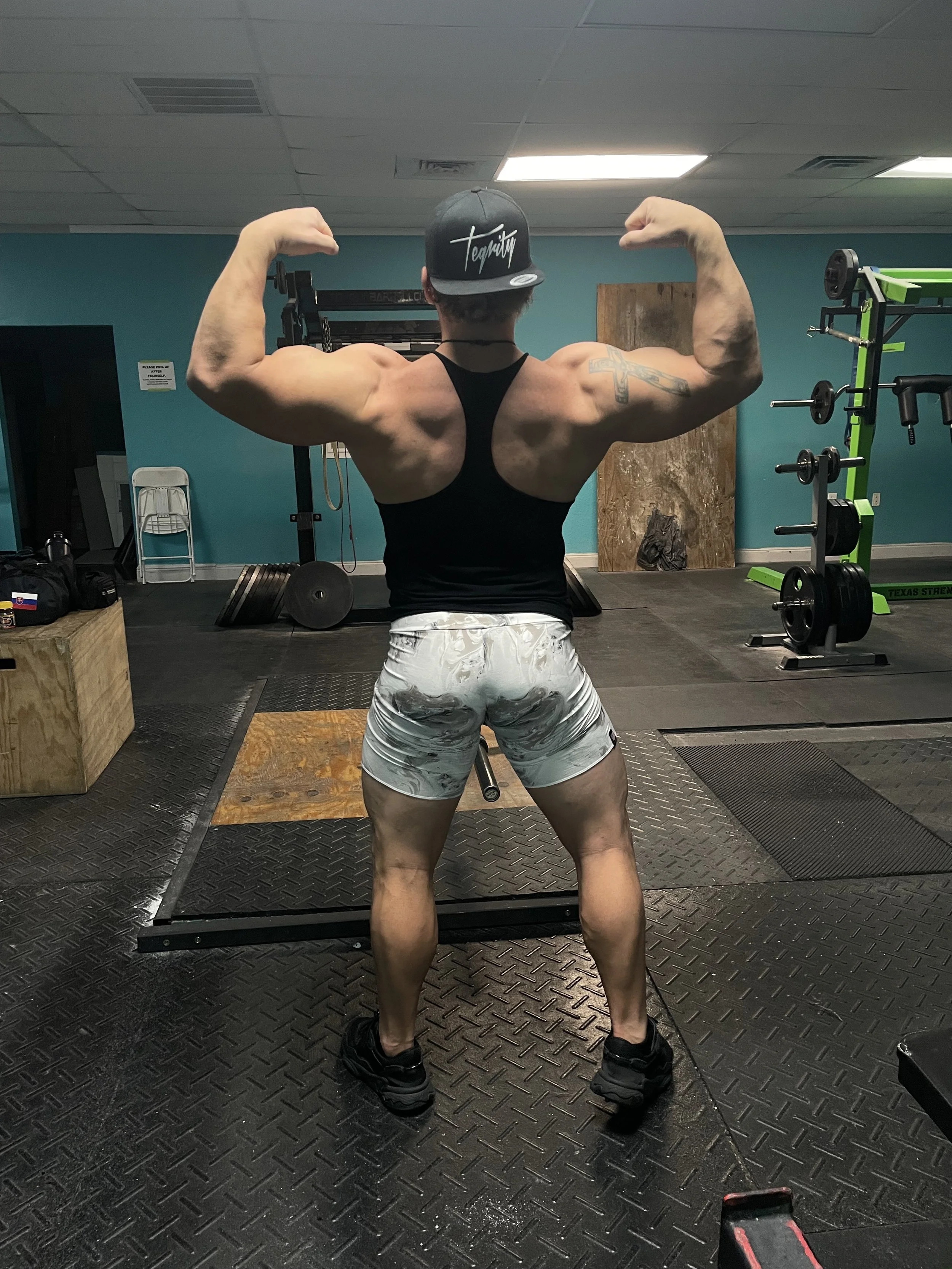 A muscular man is standing in a gym, facing away, flexing his biceps and shoulders. He is wearing a black tank top, patterned shorts, and a black cap with white writing. Gym equipment and weights are visible in the background.