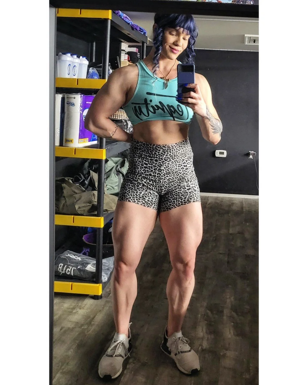 A muscular woman taking a mirror selfie in a gym, wearing a teal crop top with the word 'motivation' and leopard-print shorts, with purple hair and tattoos on her arm.