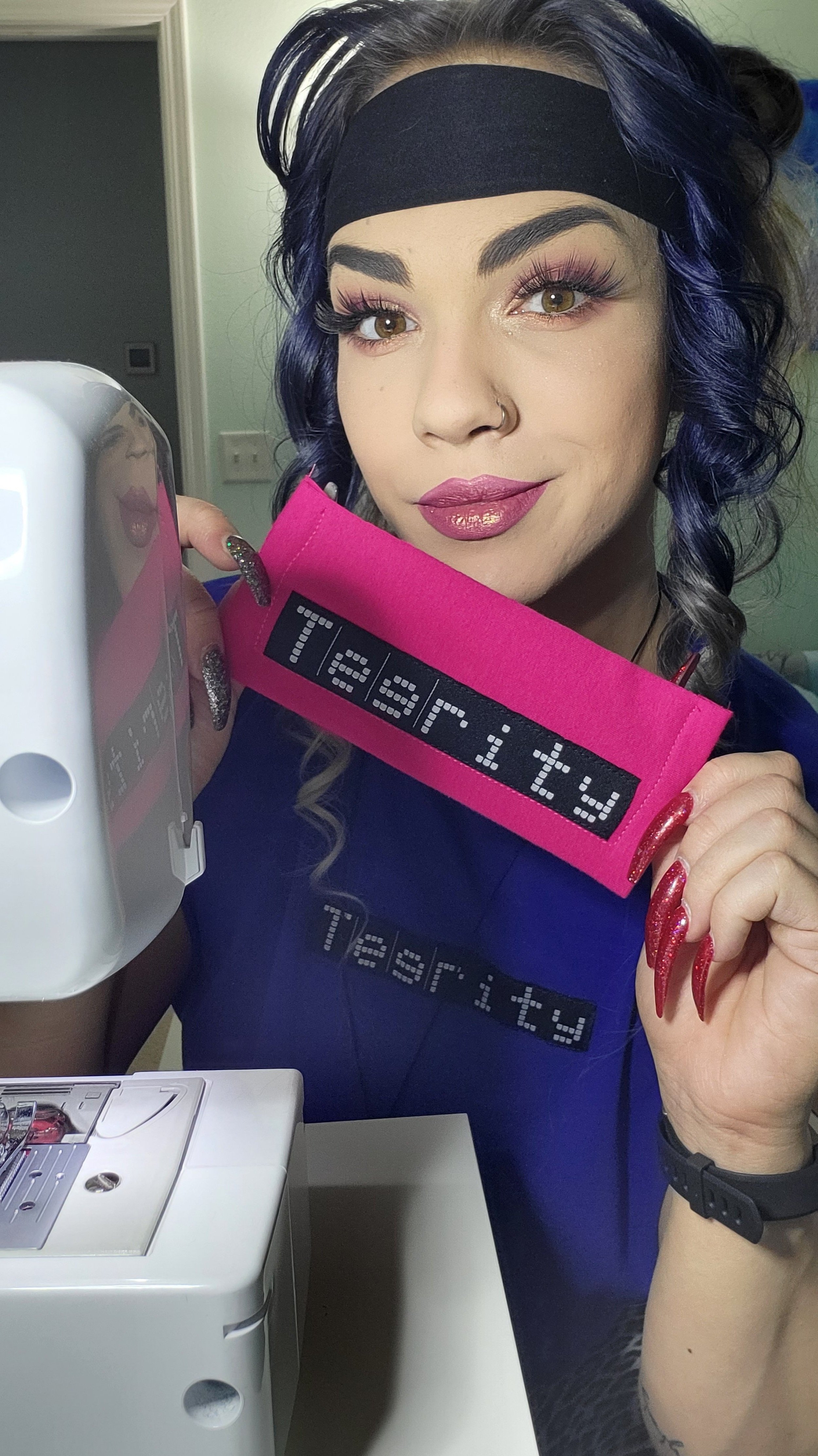 Woman with purple hair and makeup holding a pink fabric with black digital text, standing next to a sewing machine.