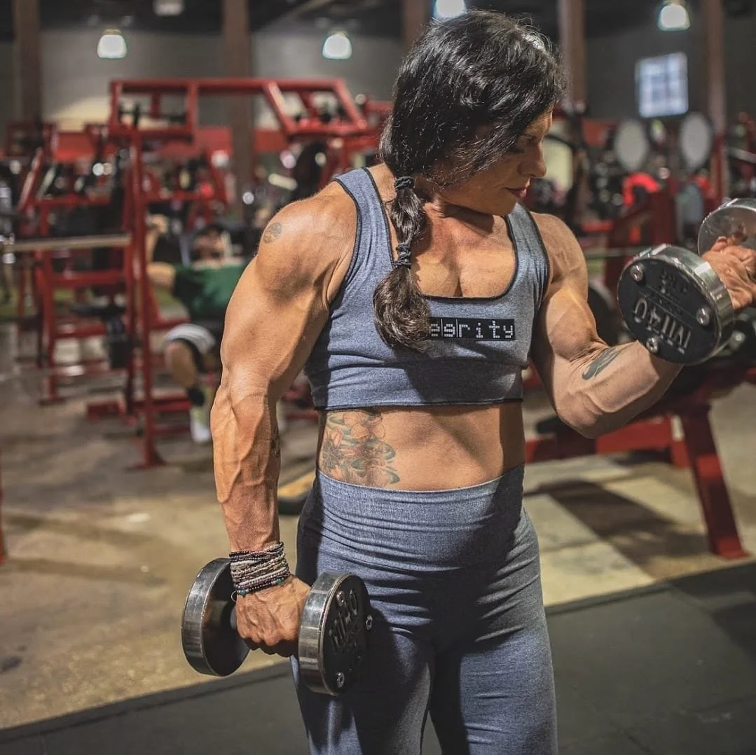 A muscular woman with a braid, wearing a sports bra with the word 'serity' on it, lifting dumbbells in a gym.