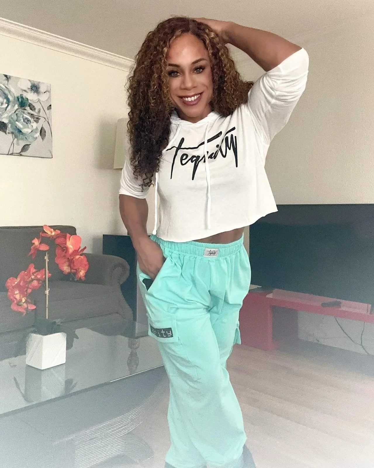 A woman with curly hair wearing a white crop hoodie and turquoise sweatpants, smiling and posing with her hand on her head in a living room with a black couch, red table, and a painting of flowers on the wall.