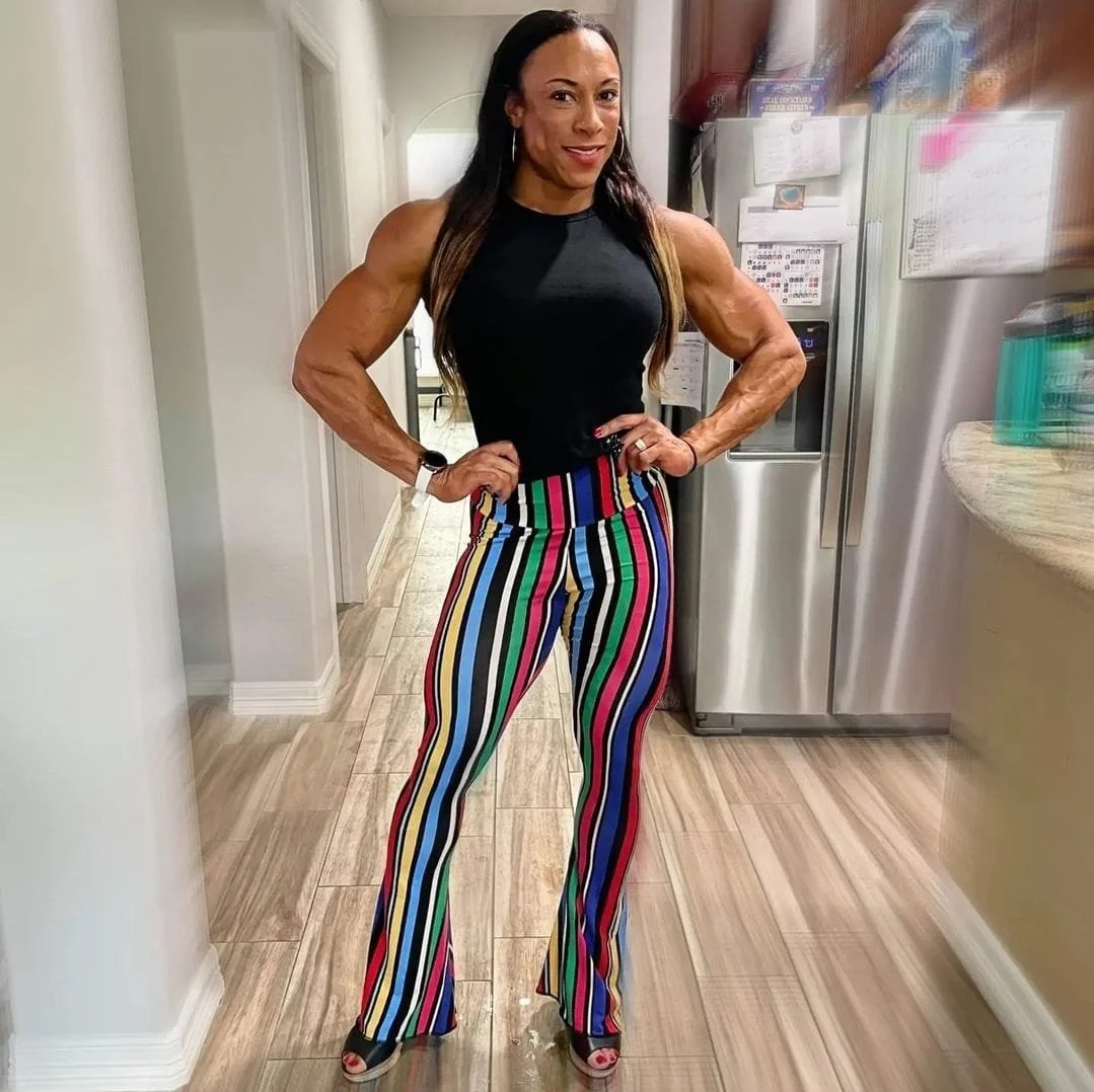 A woman standing in a kitchen with her hands on her hips, wearing a black sleeveless top and colorful striped pants.