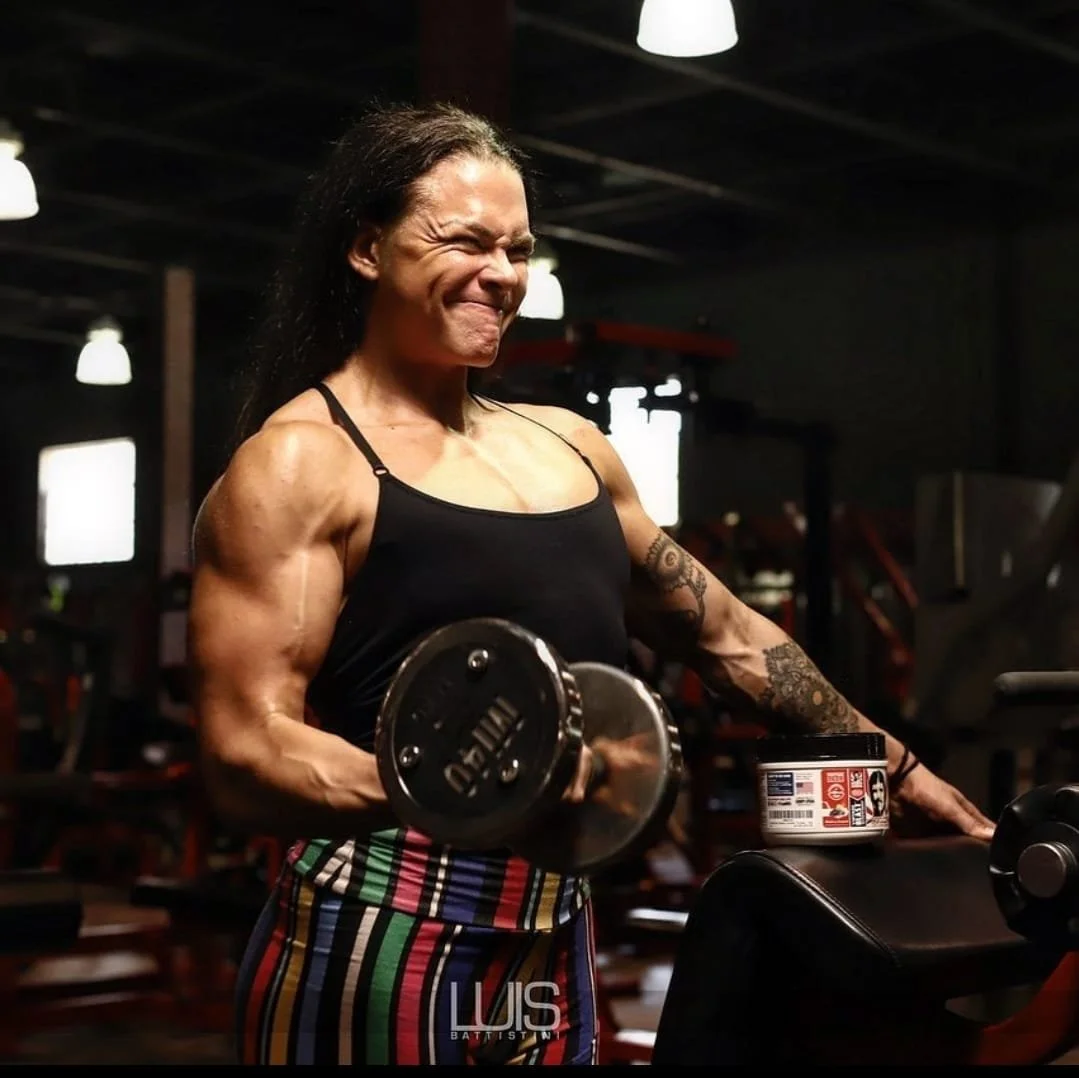 A muscular woman with long dark hair in a gym, lifting a dumbbell with a focused expression.