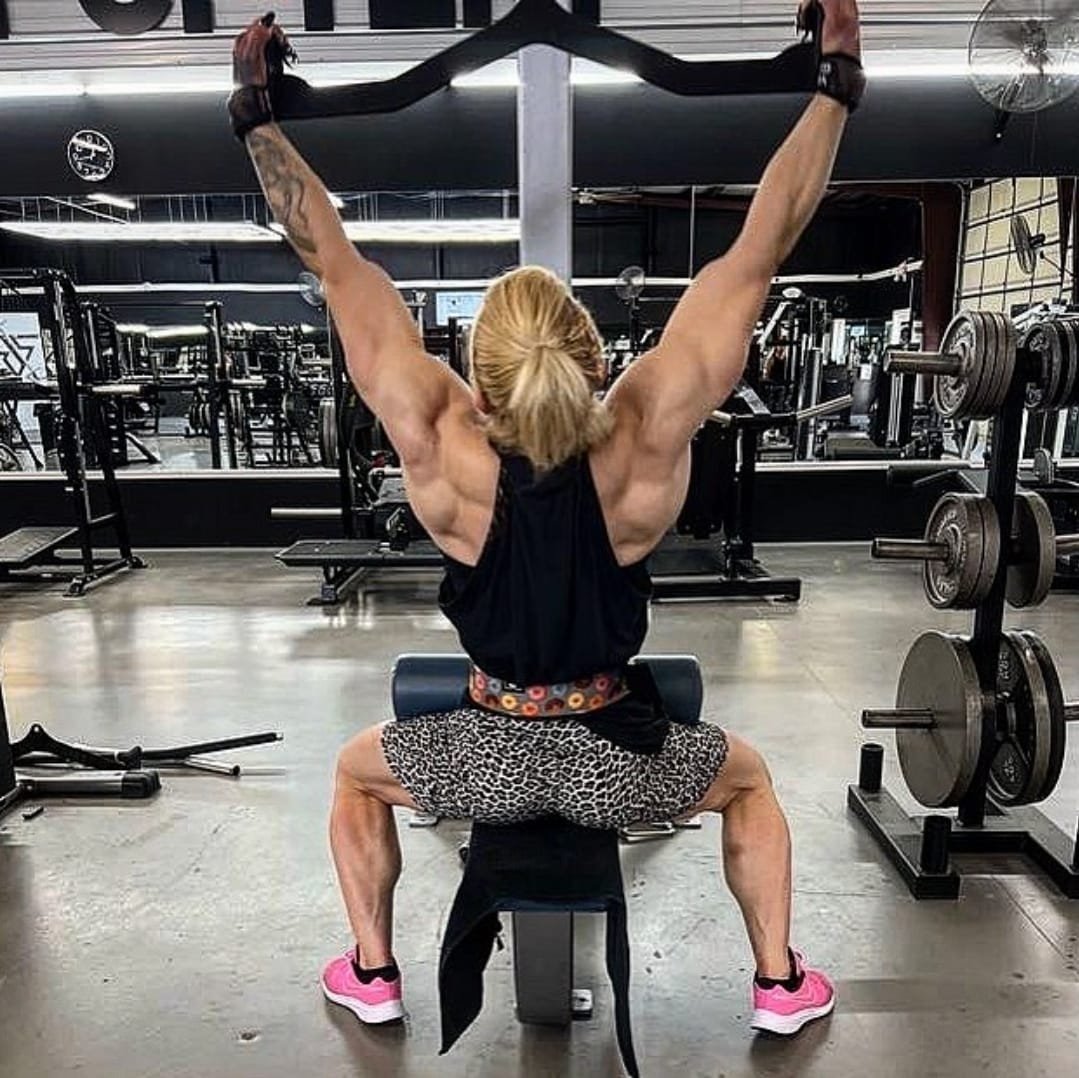A woman with muscular arms and legs sitting on a bench, holding a resistance band overhead in a gym.