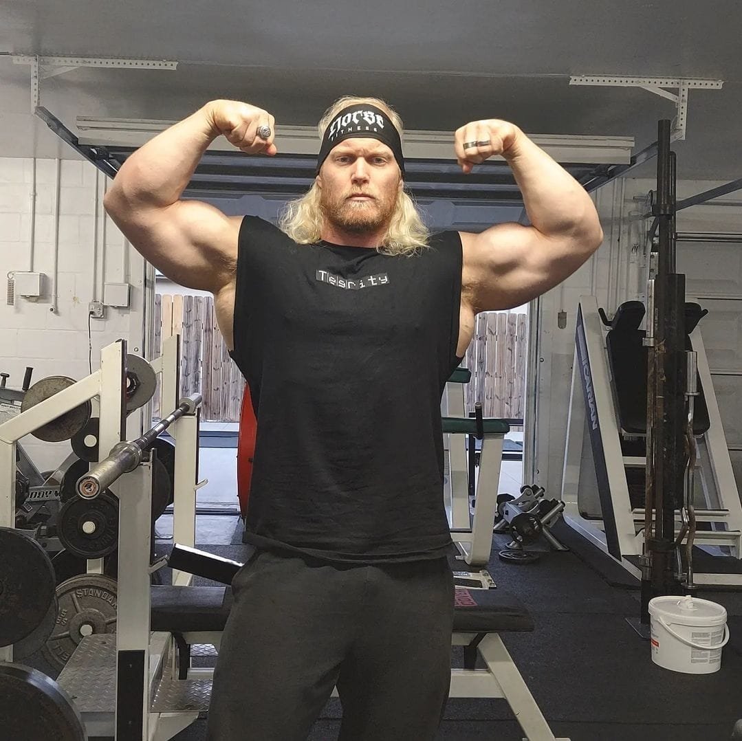 A muscular man with long blonde hair, wearing a black sleeveless shirt and headband, flexing his biceps in a gym.
