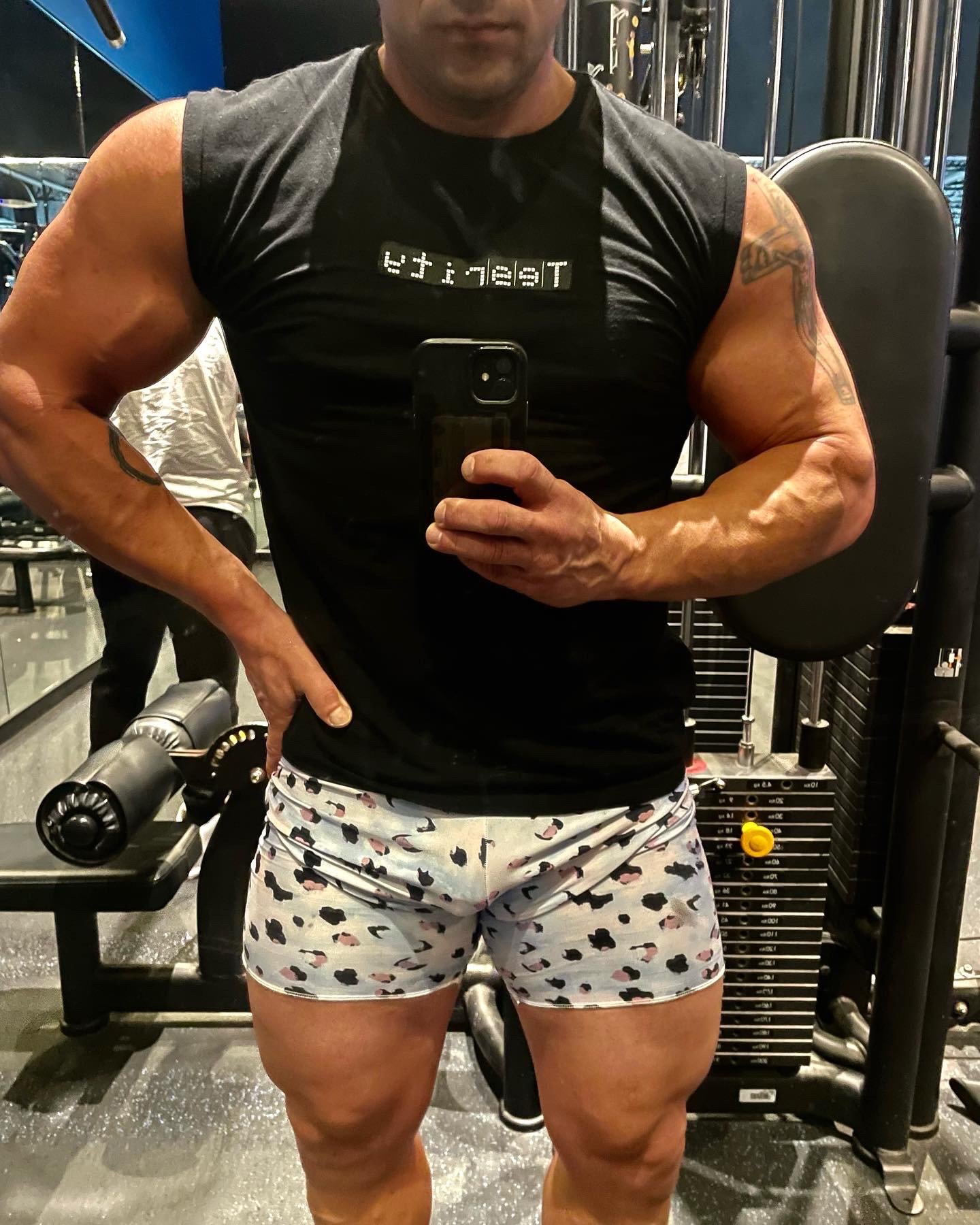 A muscular man wearing a black sleeveless shirt with the word 'Team' printed backwards and patterned shorts, taking a mirror selfie in a gym, showing his flexed arm muscles.