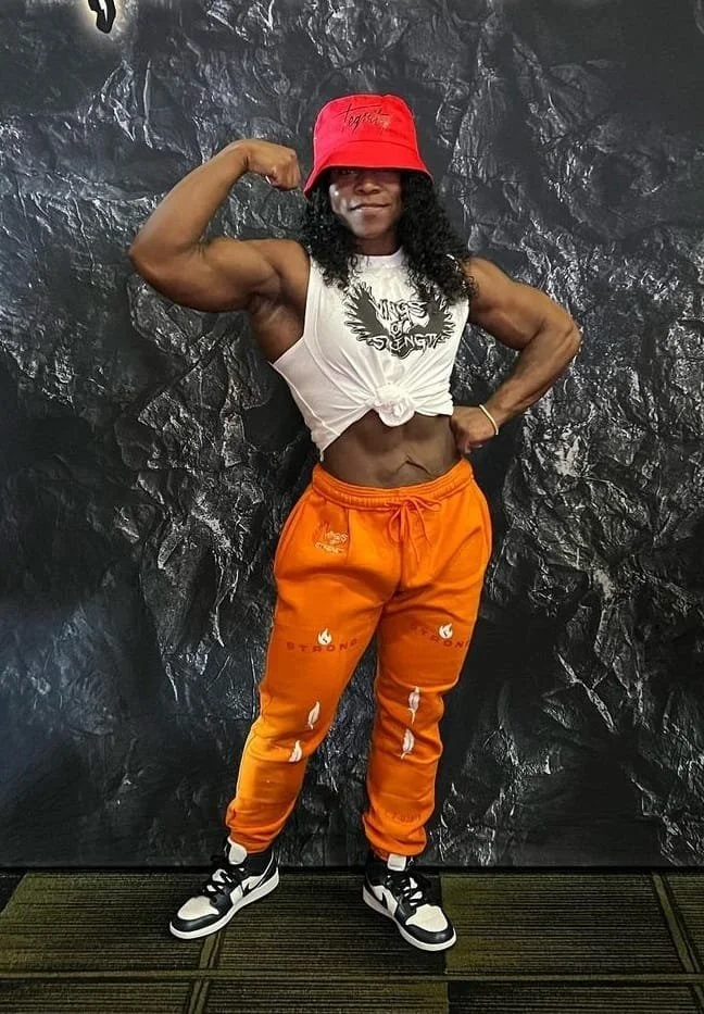 Woman flexing her arm muscles, wearing a red bucket hat, white knotted crop top, bright orange sweatpants, and black and white sneakers, standing against a dark textured background.