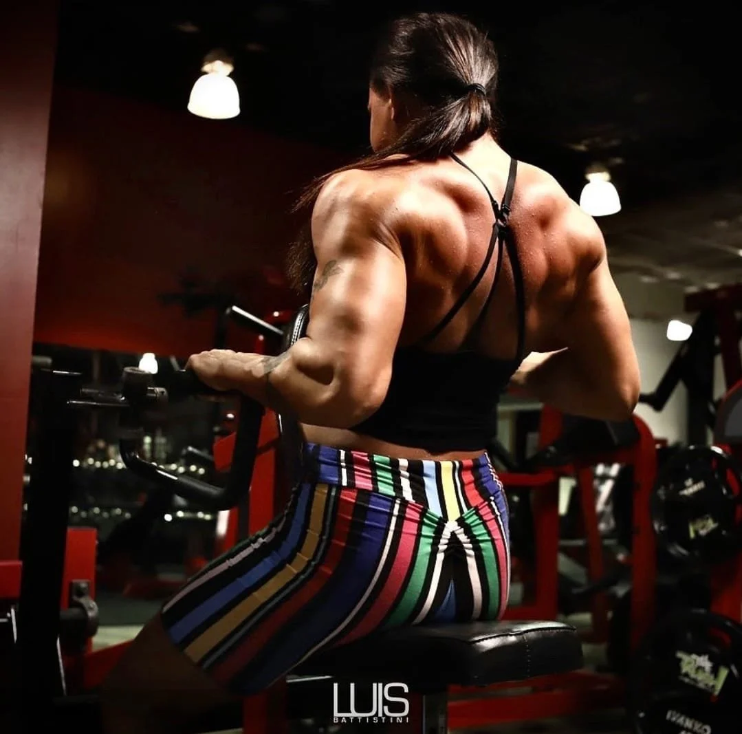 A muscular woman with tattoos sitting on a bench at the gym, viewed from behind, wearing a black strappy top and colorful vertical striped shorts, with gym equipment and mirrors in the background.