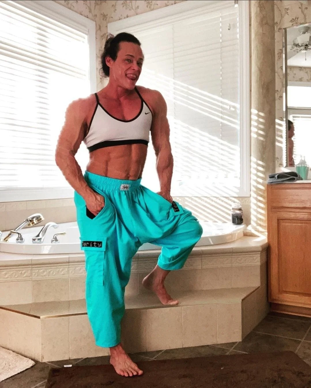 A woman with toned muscles and short dark hair posing on the edge of a bathtub in a bathroom, wearing a white sports bra and bright turquoise pants, with sunlight streaming through window blinds.