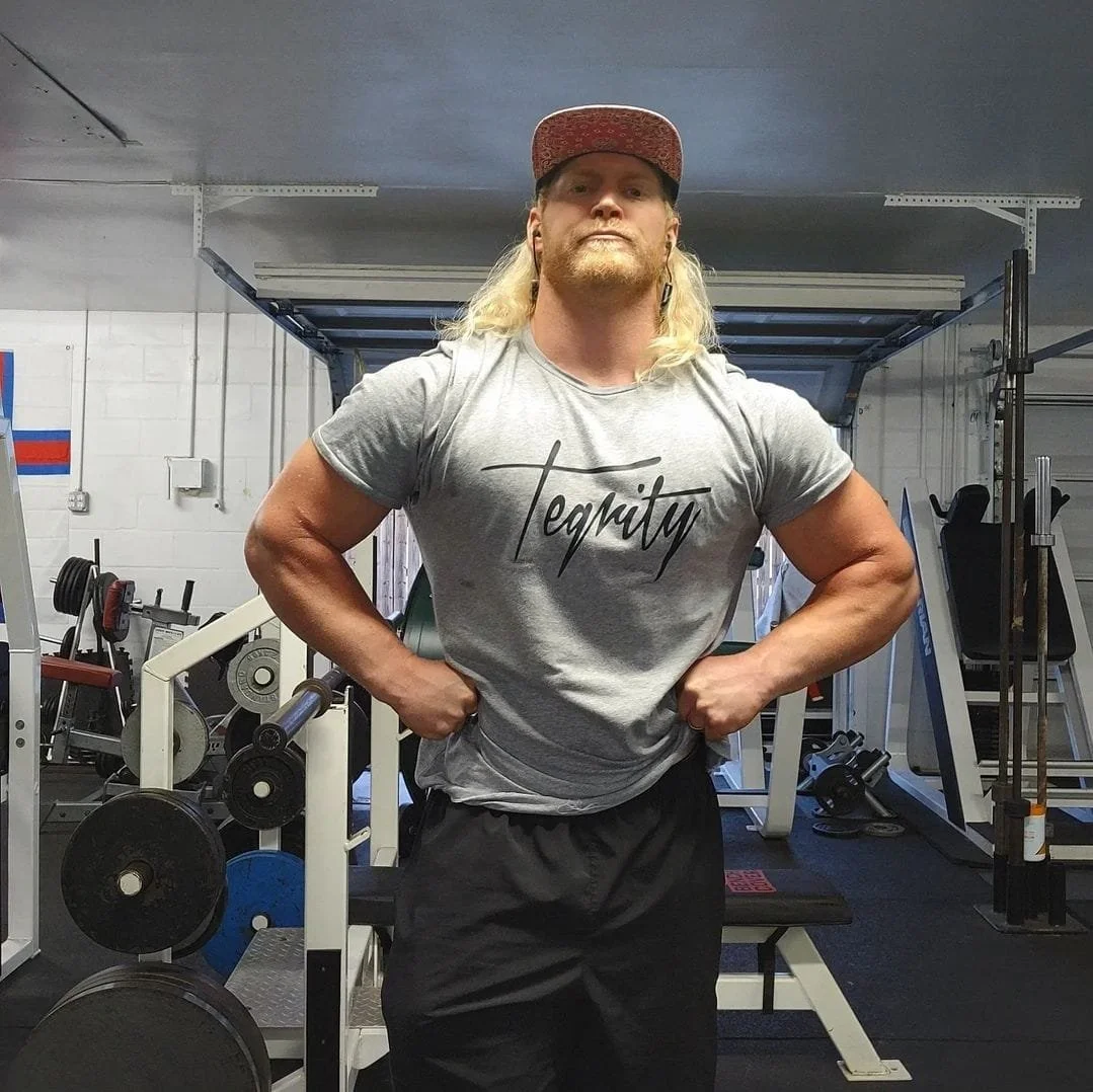 A muscular man with long blond hair, wearing a red cap and a gray t-shirt with the word 'Tolerance' written on it, standing with hands on hips in a gym.
