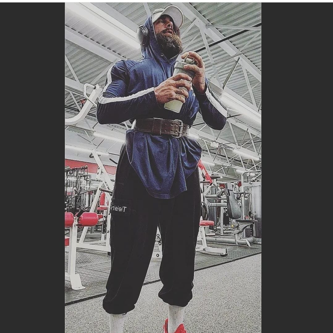 A man with a beard at the gym wearing a hoodie, headphones, and sweatpants holding a water bottle, standing in front of gym equipment.