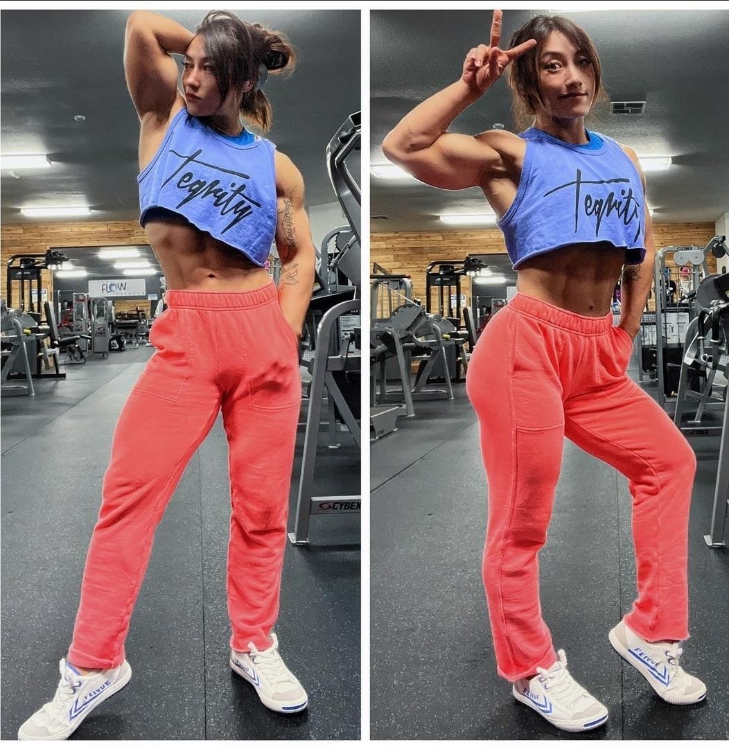A woman at the gym posing in front of workout equipment, wearing a blue cropped tank top with 'Tenacity' written on it, red sweatpants, and white sneakers.