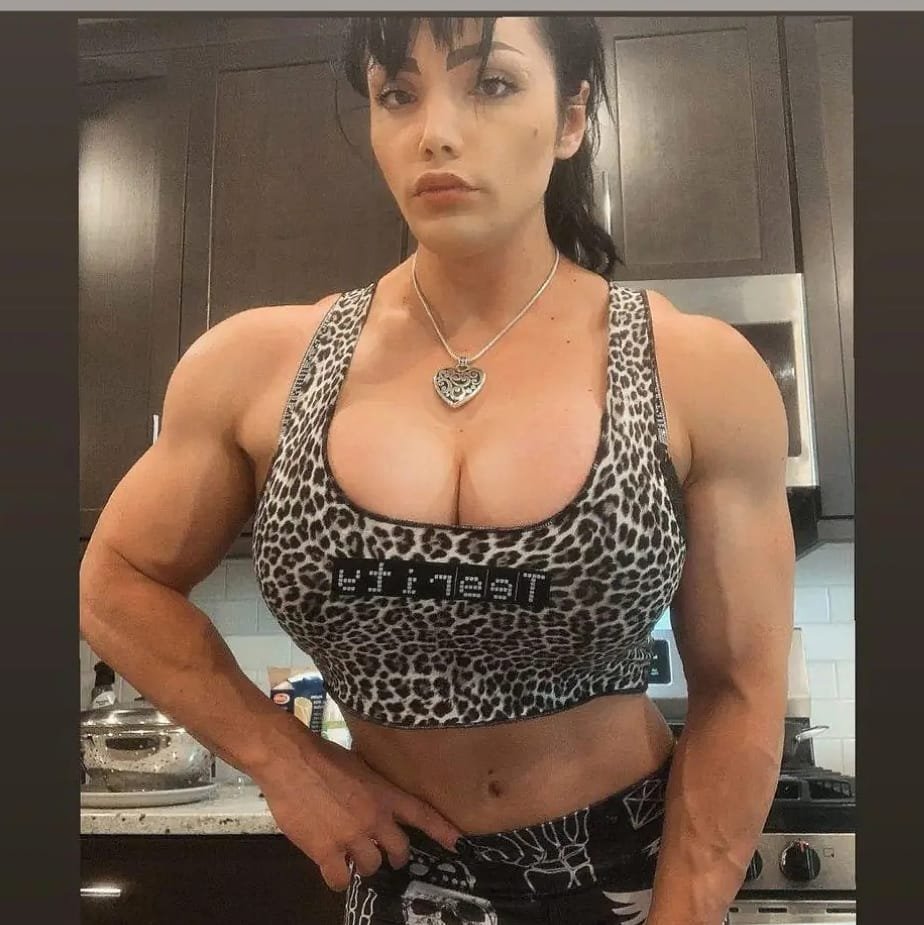 A muscular woman with short dark hair in a kitchen, wearing a leopard print crop top and black pants with spider web designs, posing with her hand on her waist.