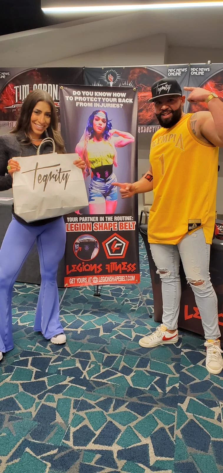 A woman and a man posing at a health and fitness event, with a banner promoting Legion Shape Belt and fitness products in the background.