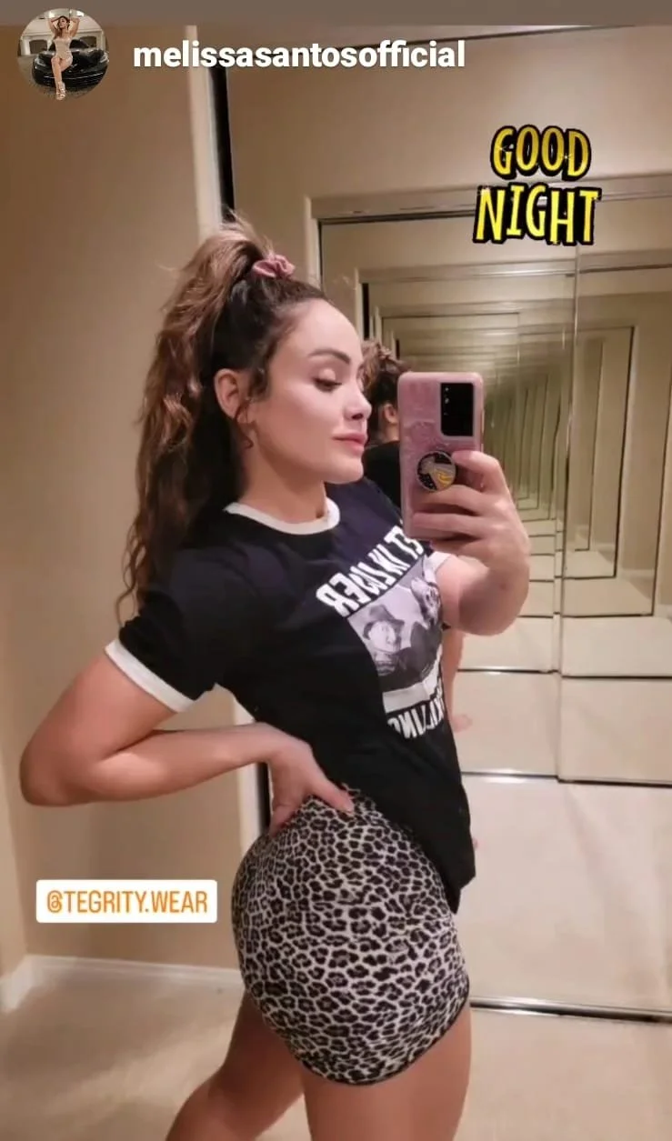 A young woman taking a mirror selfie in a hall with mirrored walls, wearing a graphic t-shirt and leopard print shorts, with her hair in a high ponytail. The image has a 'Good Night' sticker and an Instagram handle '@TEGRITY.WEAR'.