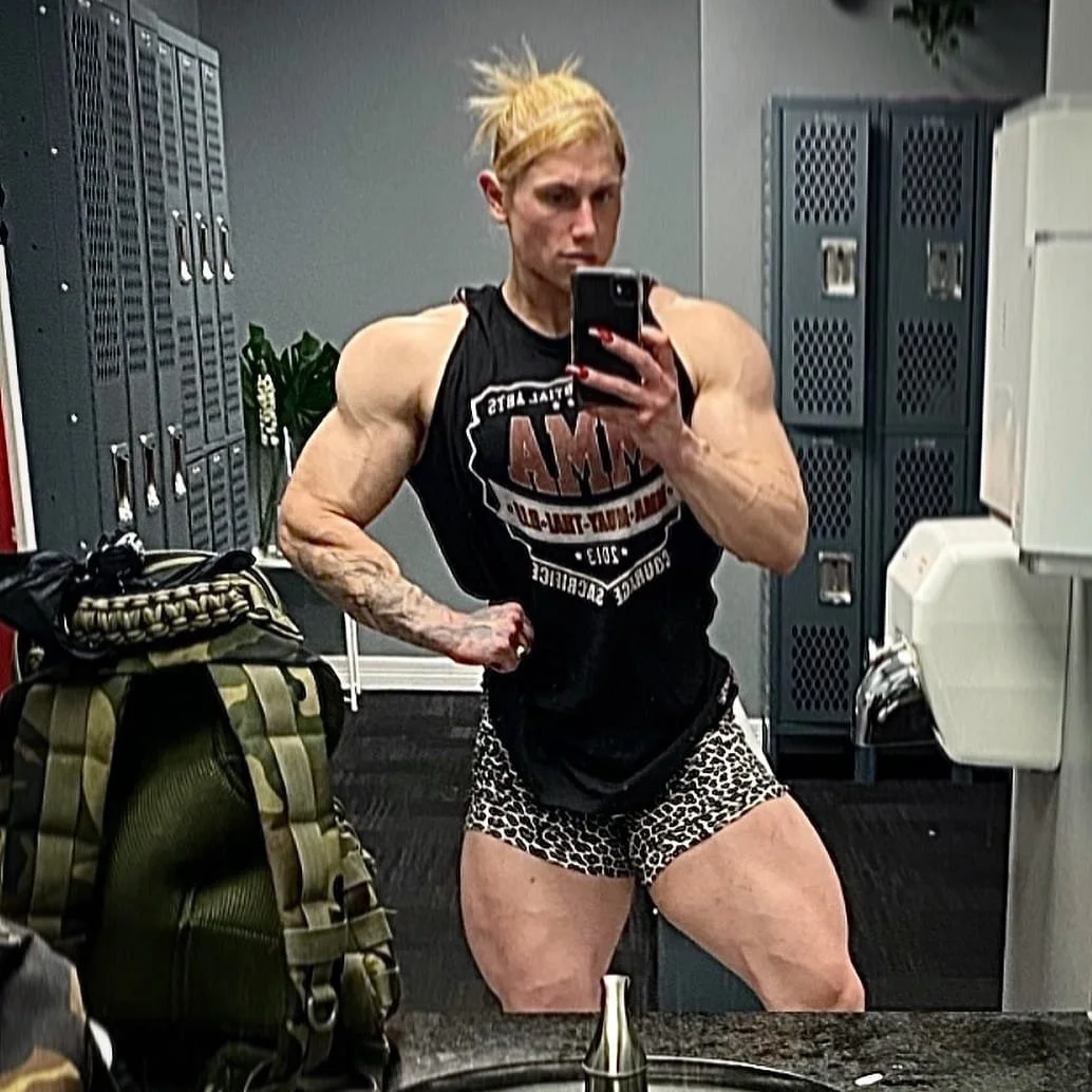 A muscular woman with blond hair in a messy bun taking a selfie in a gym locker room mirror, showcasing her defined arms and legs, wearing a black sleeveless shirt and leopard print shorts.