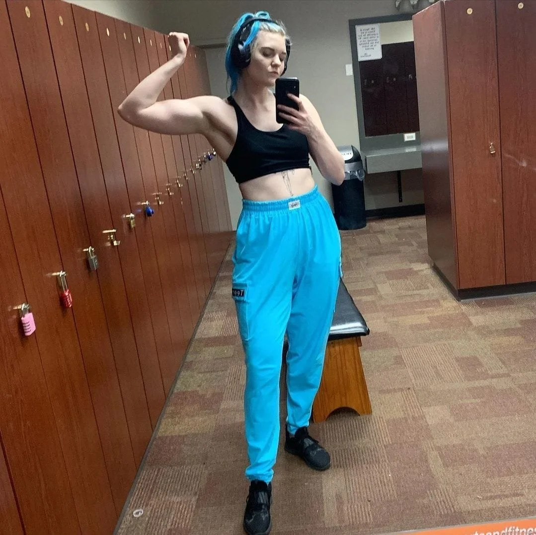A young woman takes a selfie in a gym locker room, flexing her right arm to showcase her bicep. She has dyed blue hair, wears a black sports bra, bright blue joggers, and black sneakers, with large blue over-ear headphones.