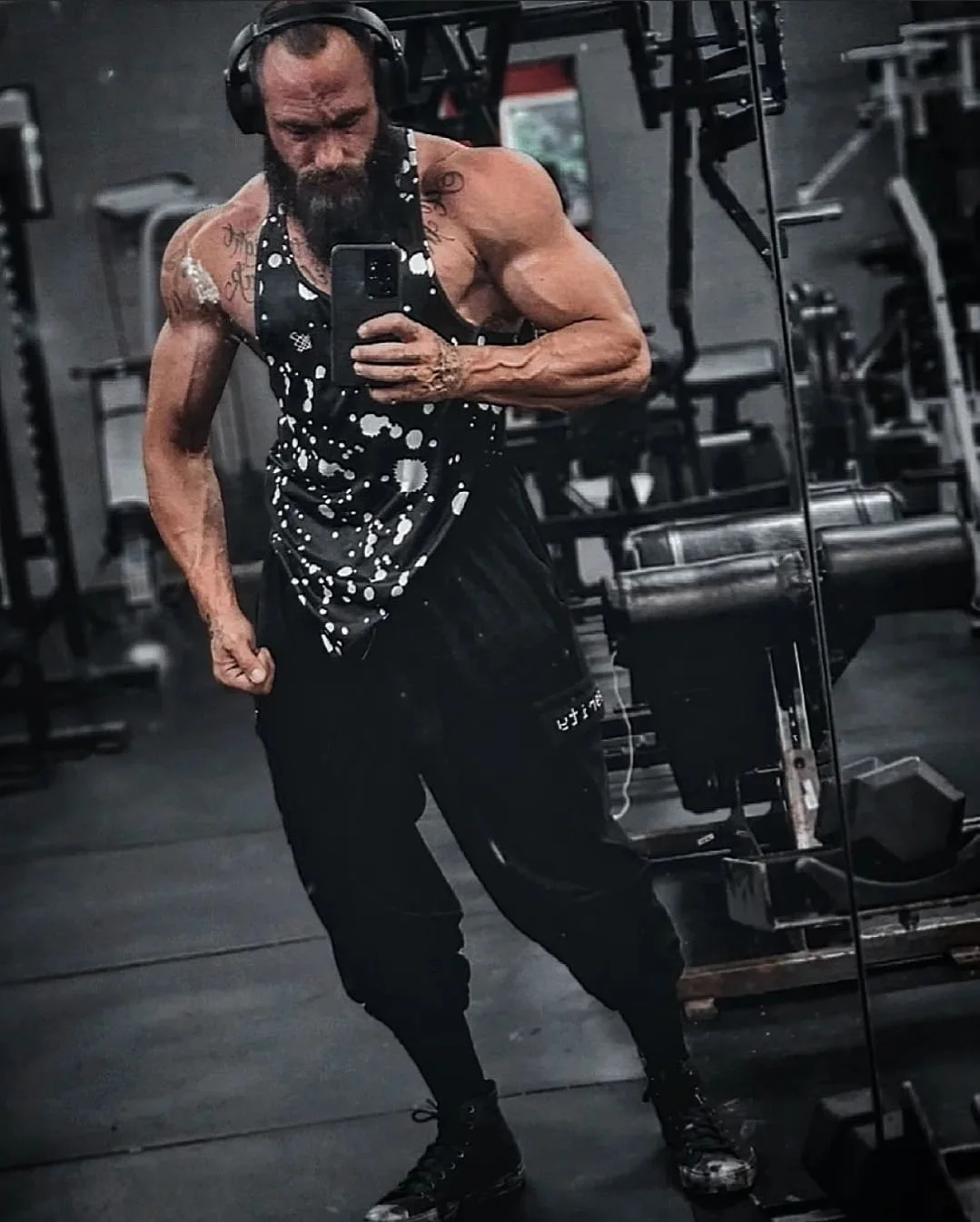 A muscular man taking a mirror selfie in a gym, wearing a black and white splatter-pattern tank top, black pants, and headphones.