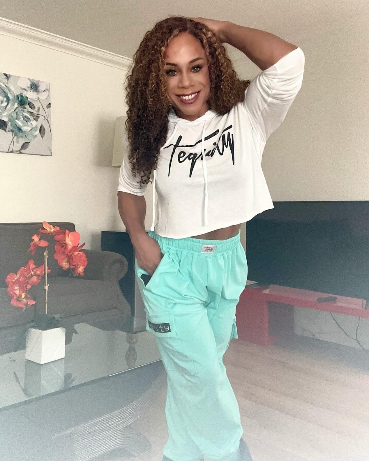 A smiling woman with curly hair wearing a white hoodie with black lettering and light green sweatpants, standing in a living room.