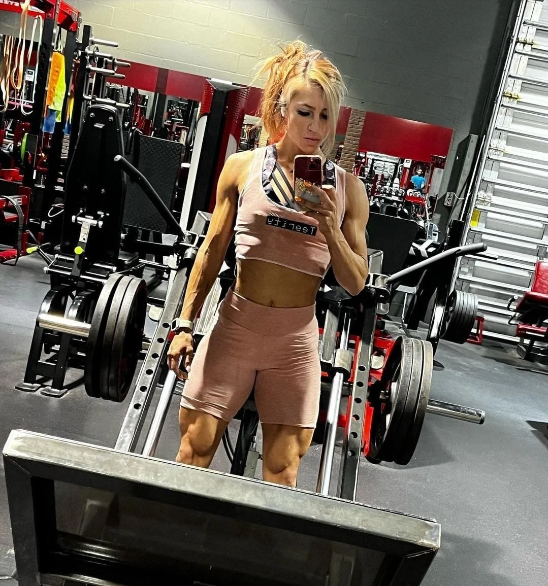 A woman taking a mirror selfie in a gym, wearing a pink sports bra and matching shorts, with various workout equipment around her.