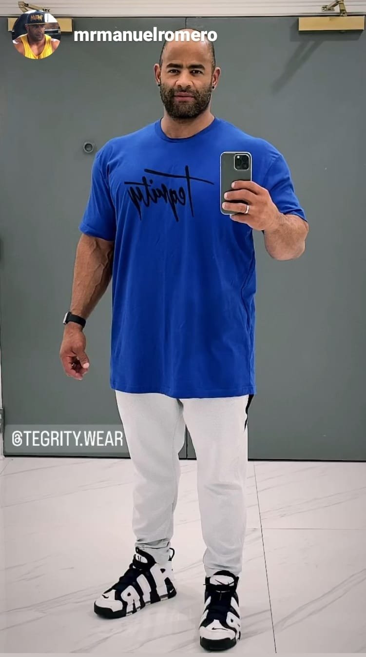 A man with a beard is taking a selfie in front of a mirror, wearing a blue 'Dolphin' t-shirt, white pants, and black-and-white sneakers with a bold pattern, in a room with a gray wall and tiled floor.